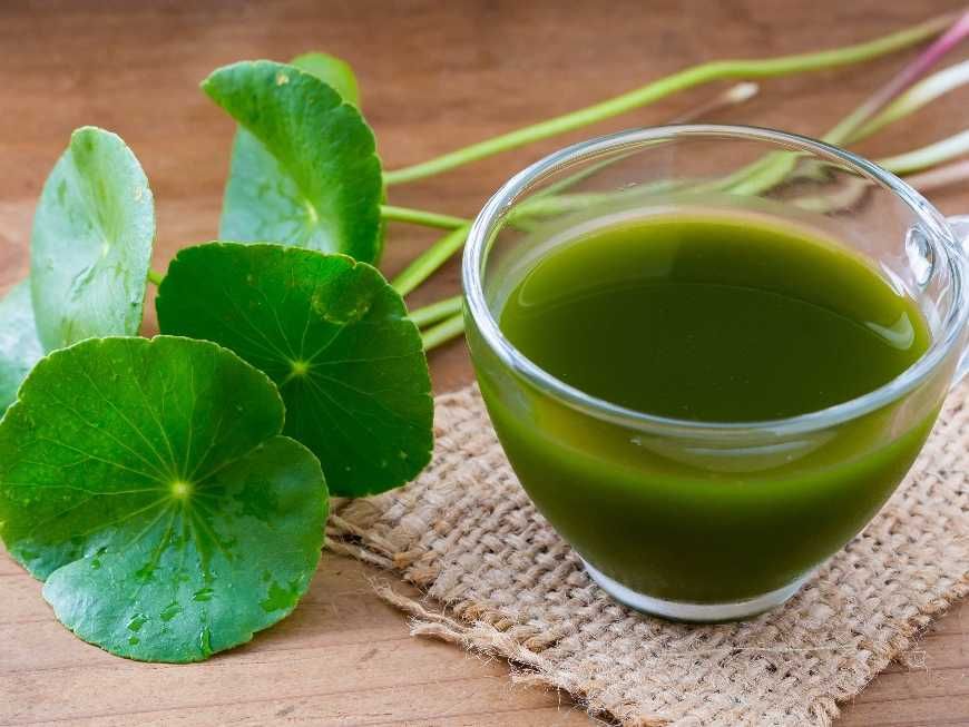 Green herbal drink in a glass cup, next to green leaves on a wood surface.