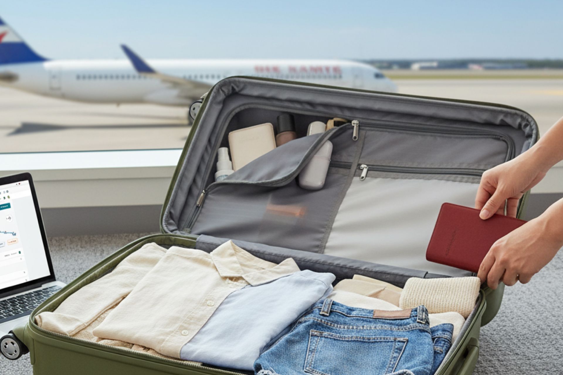 Person packing suitcase at airport, near laptop and plane. Hand holding passport; clothes neatly arranged.