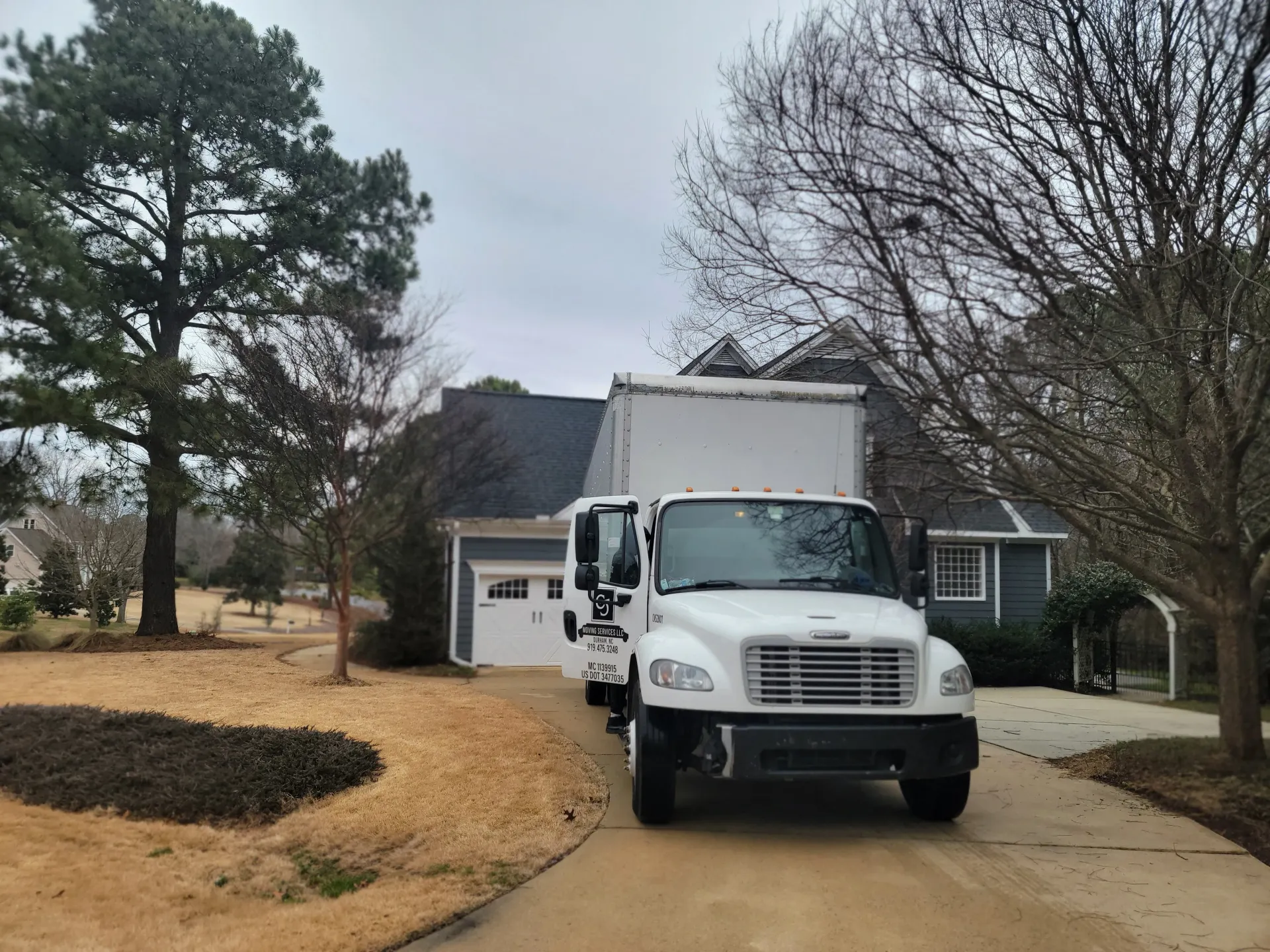 A white box truck parked in the driveway of a residential house on a cloudy day with bare trees in the yard.