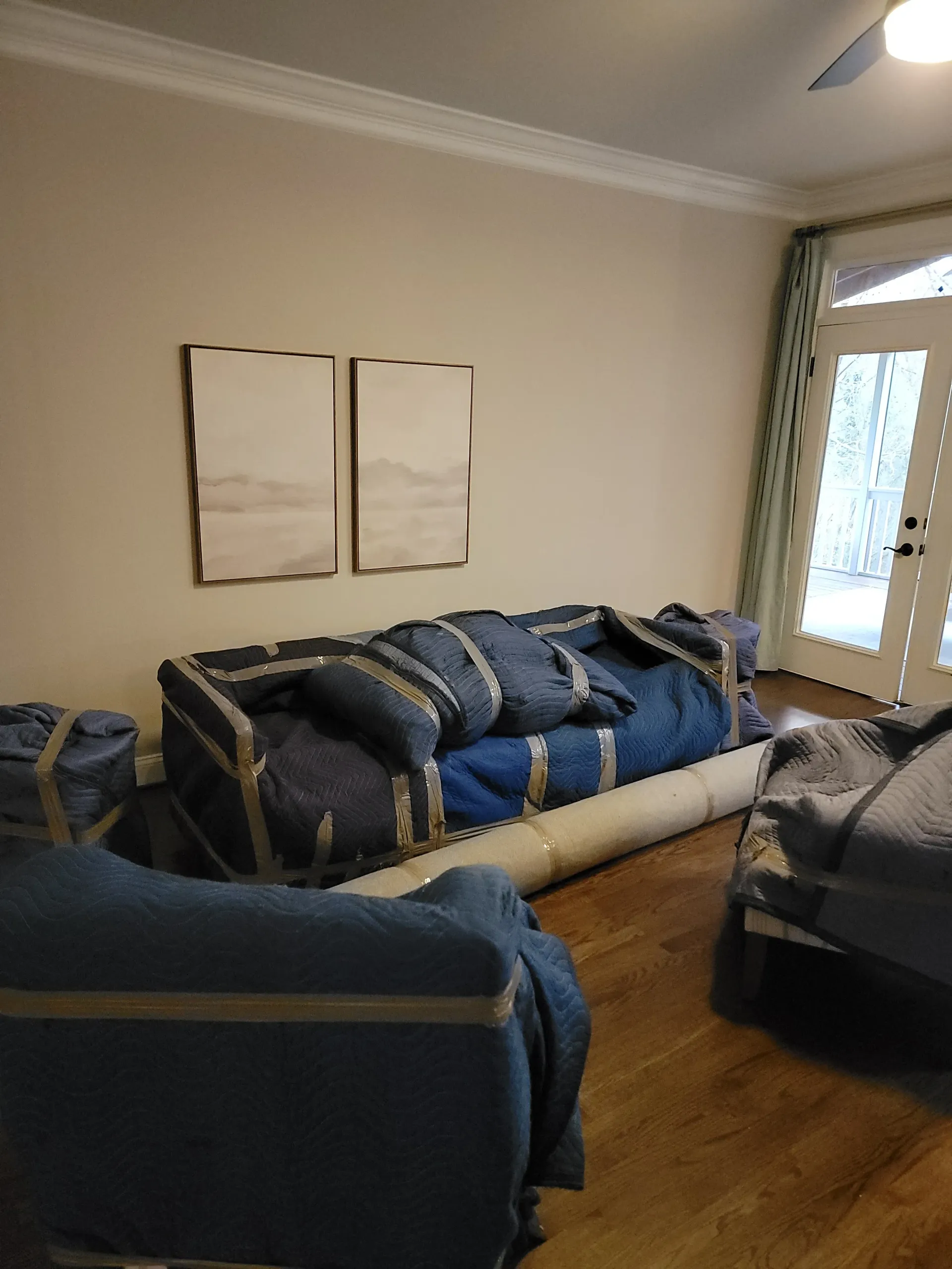 Furniture wrapped in blue moving blankets sits on a hardwood floor in a room with neutral walls and two framed pictures.