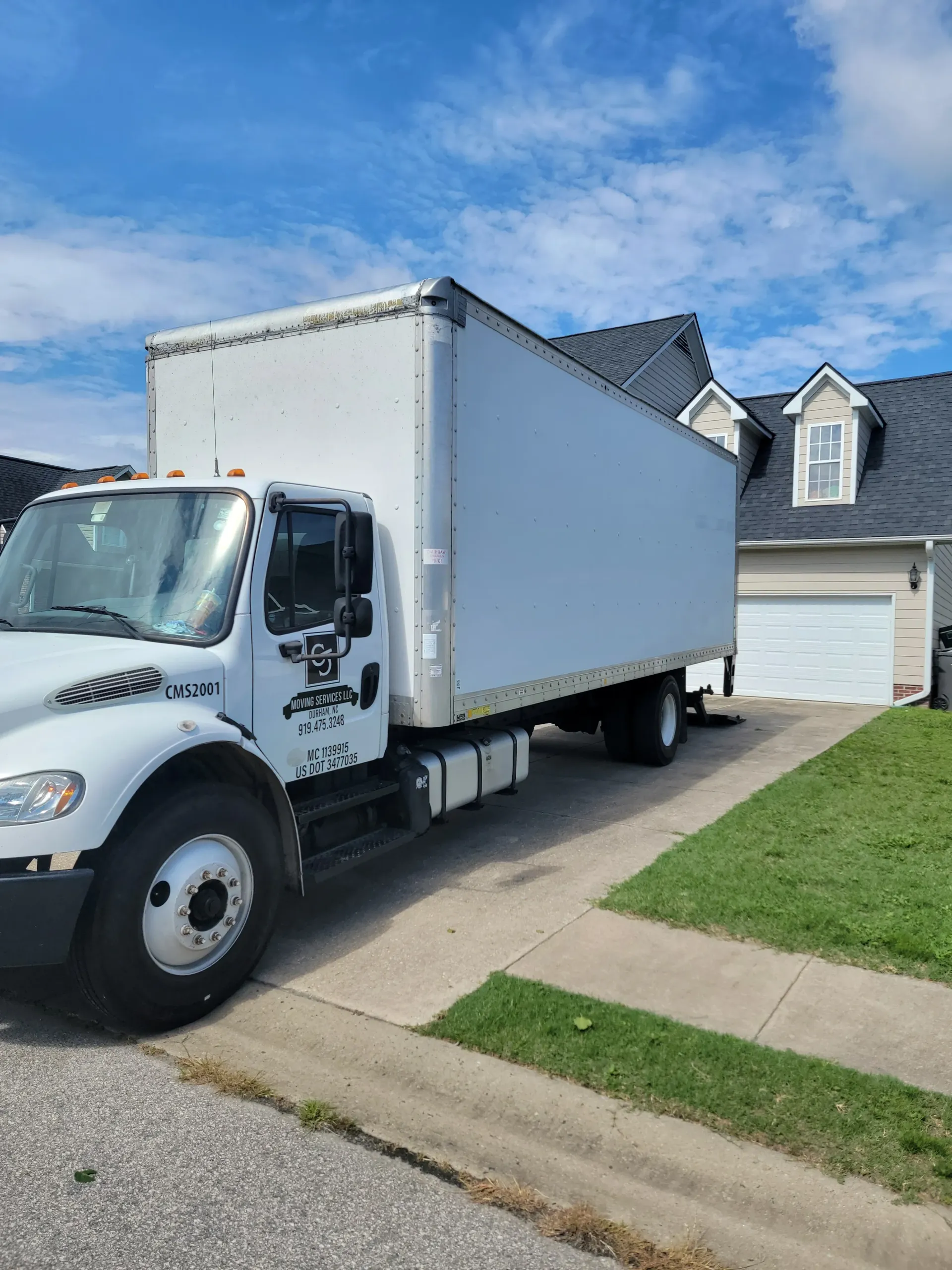 A large white box truck parked in a residential driveway in front of a house with a garage.