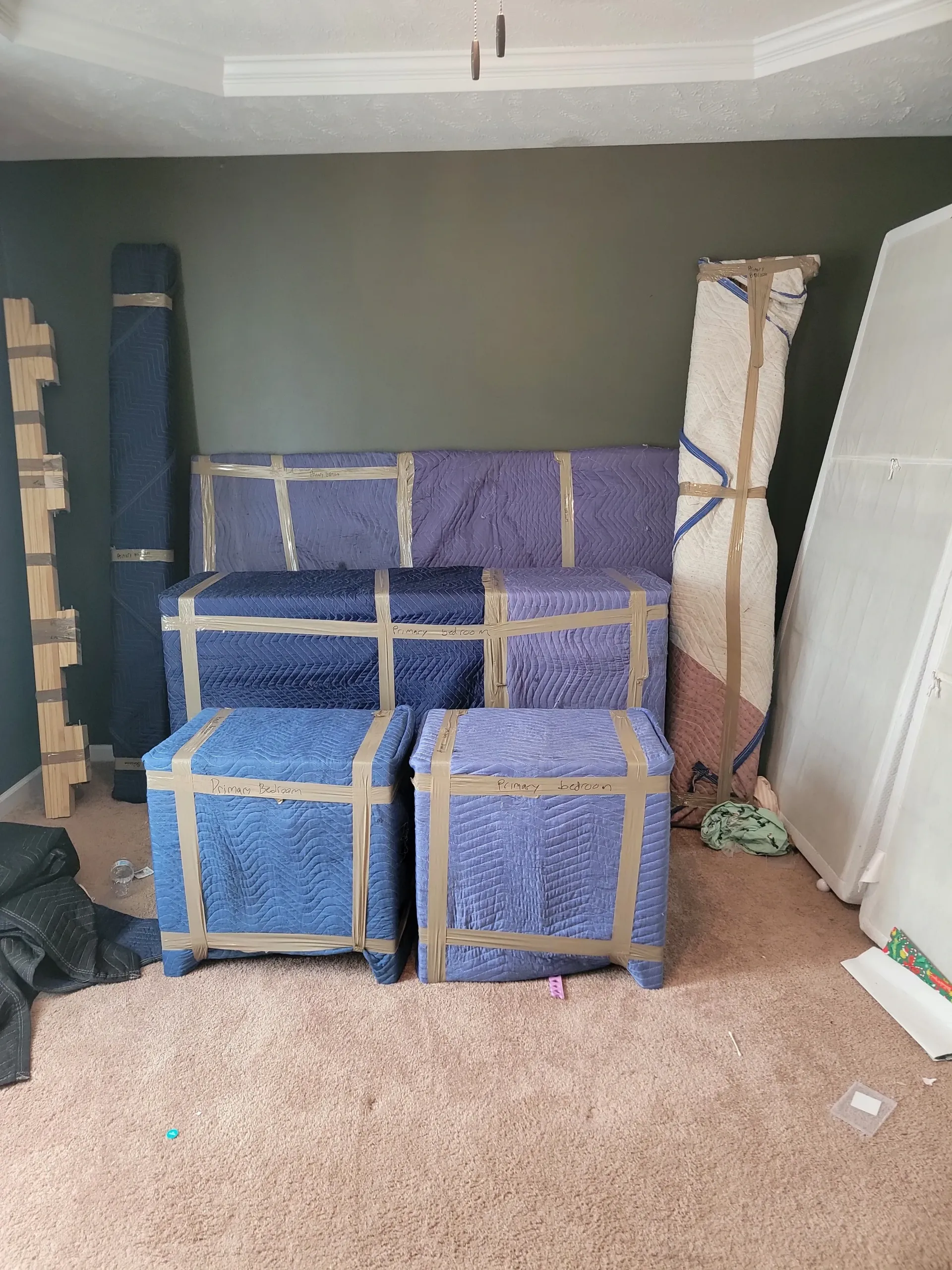 Furniture items wrapped in blue protective blankets and secured with packing tape inside a room with a dark accent wall.