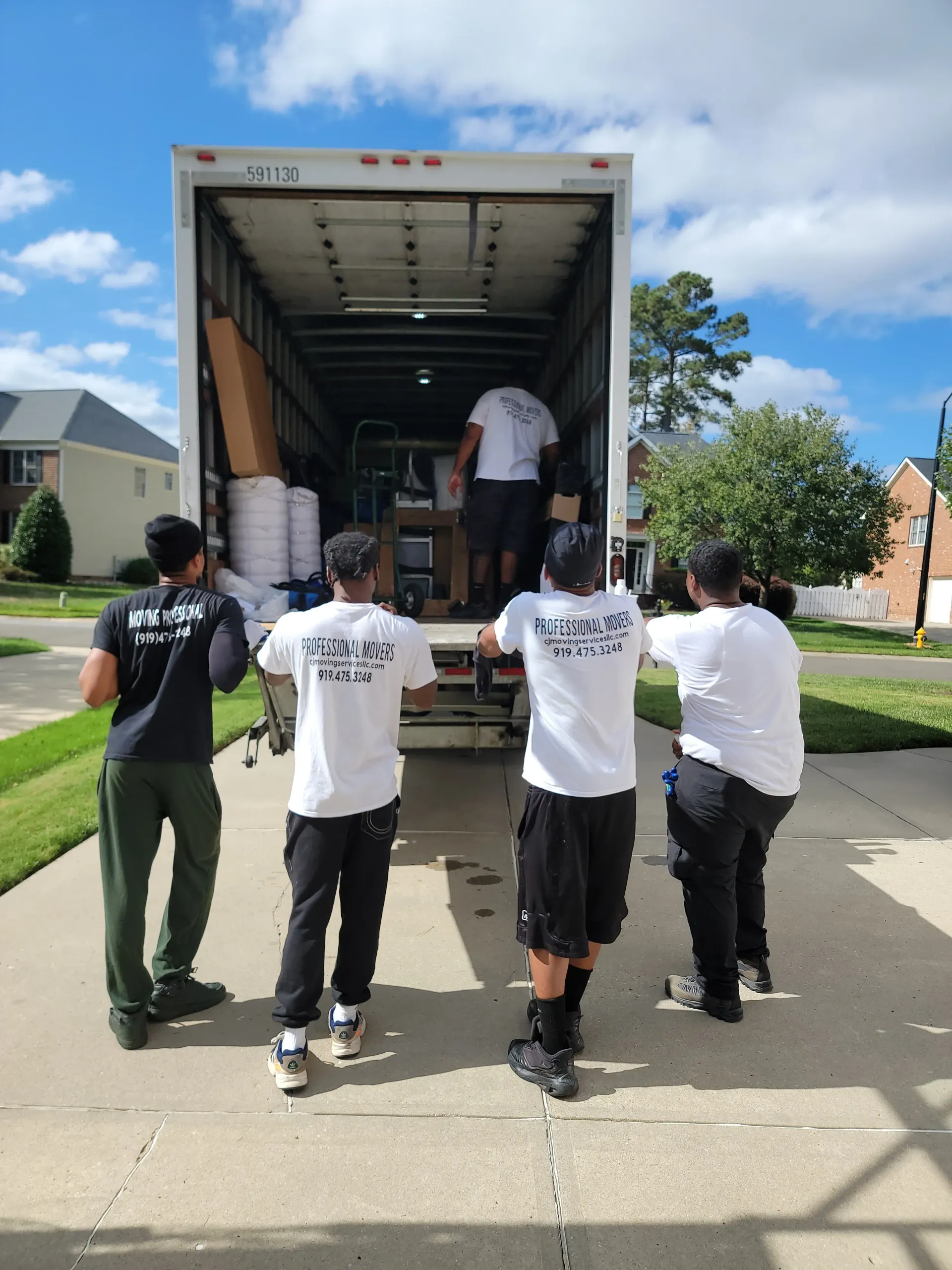 Five people in matching shirts stand behind a large moving truck parked on a suburban driveway, preparing to unload items.