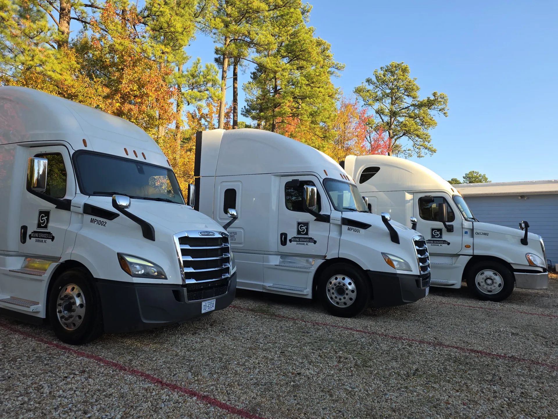 Three identical white semi-trucks parked in a row on a gravel lot in front of trees under a clear blue sky.