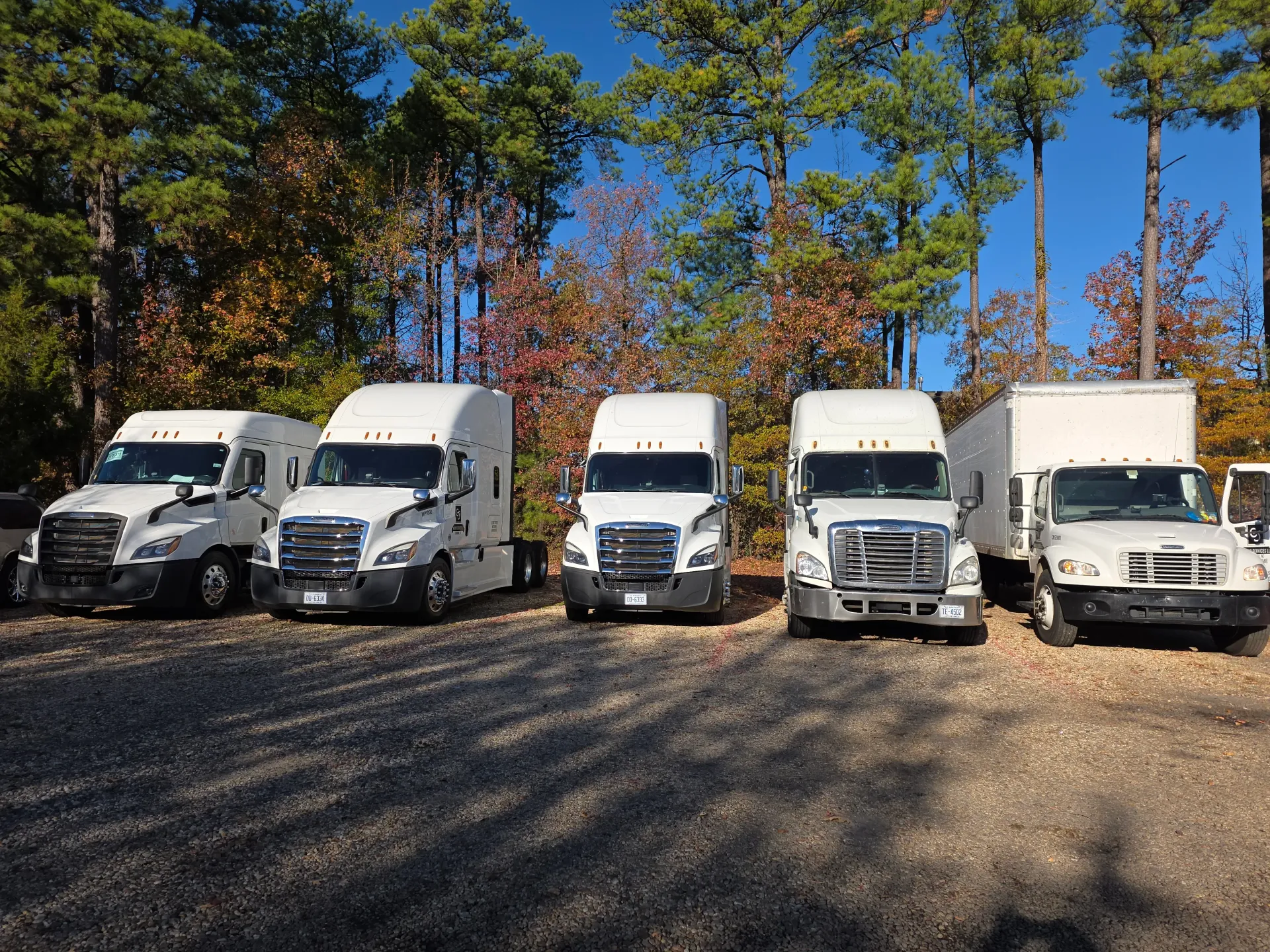 A row of five white semi-trucks and delivery vehicles parked on a gravel lot in front of autumn-colored trees.