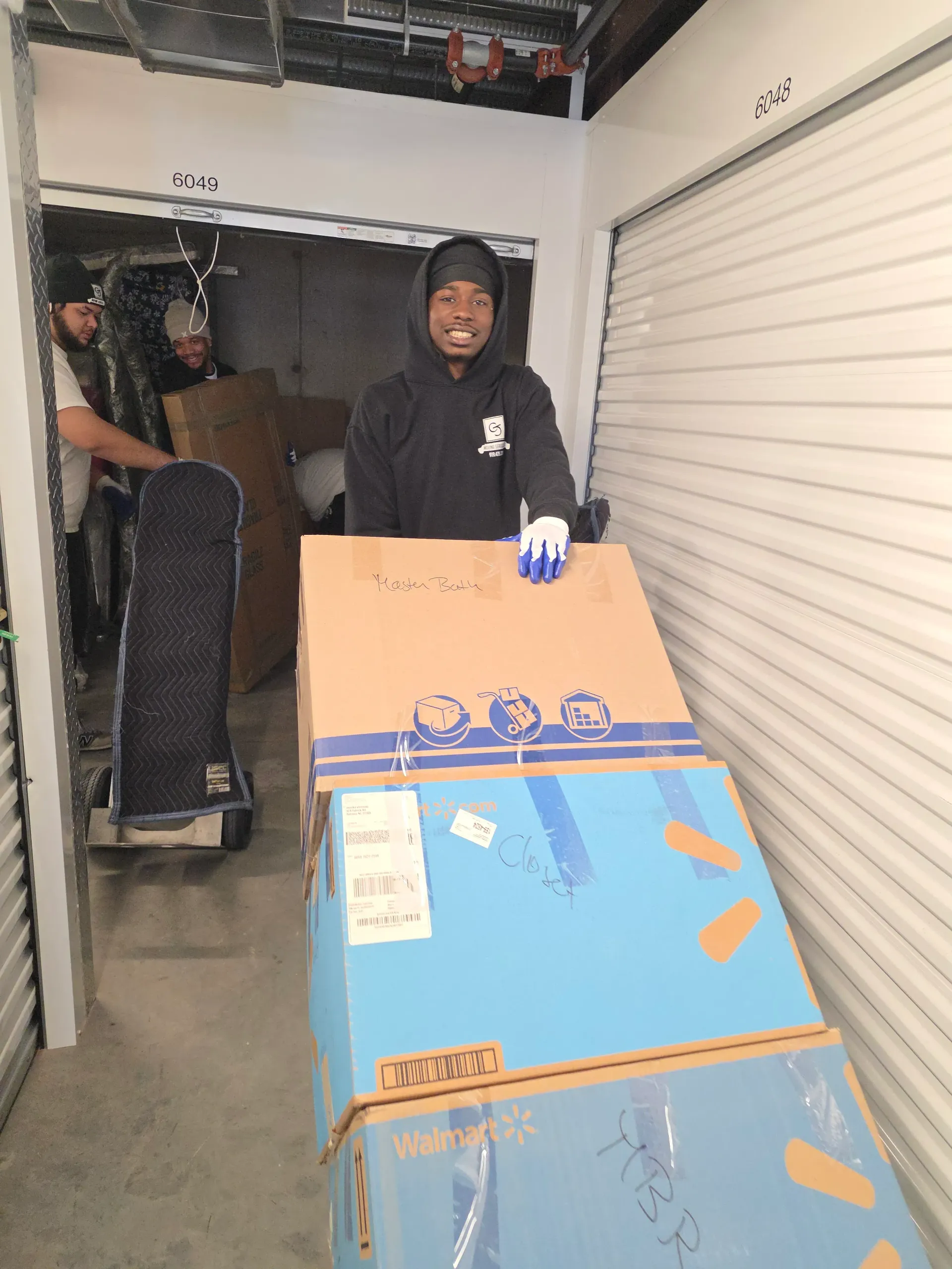 A person wearing a hoodie and work gloves uses a hand truck to move stacked boxes inside a storage unit.
