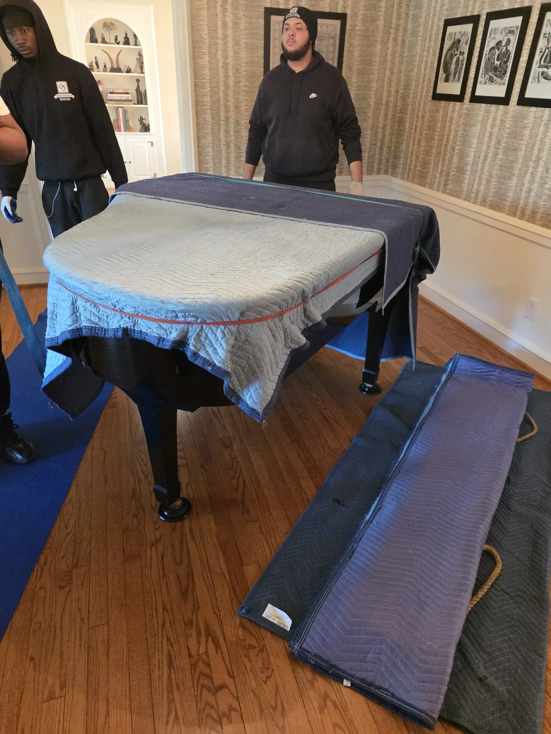 Two people in hooded sweatshirts stand by a grand piano covered with protective moving blankets in a residential room.