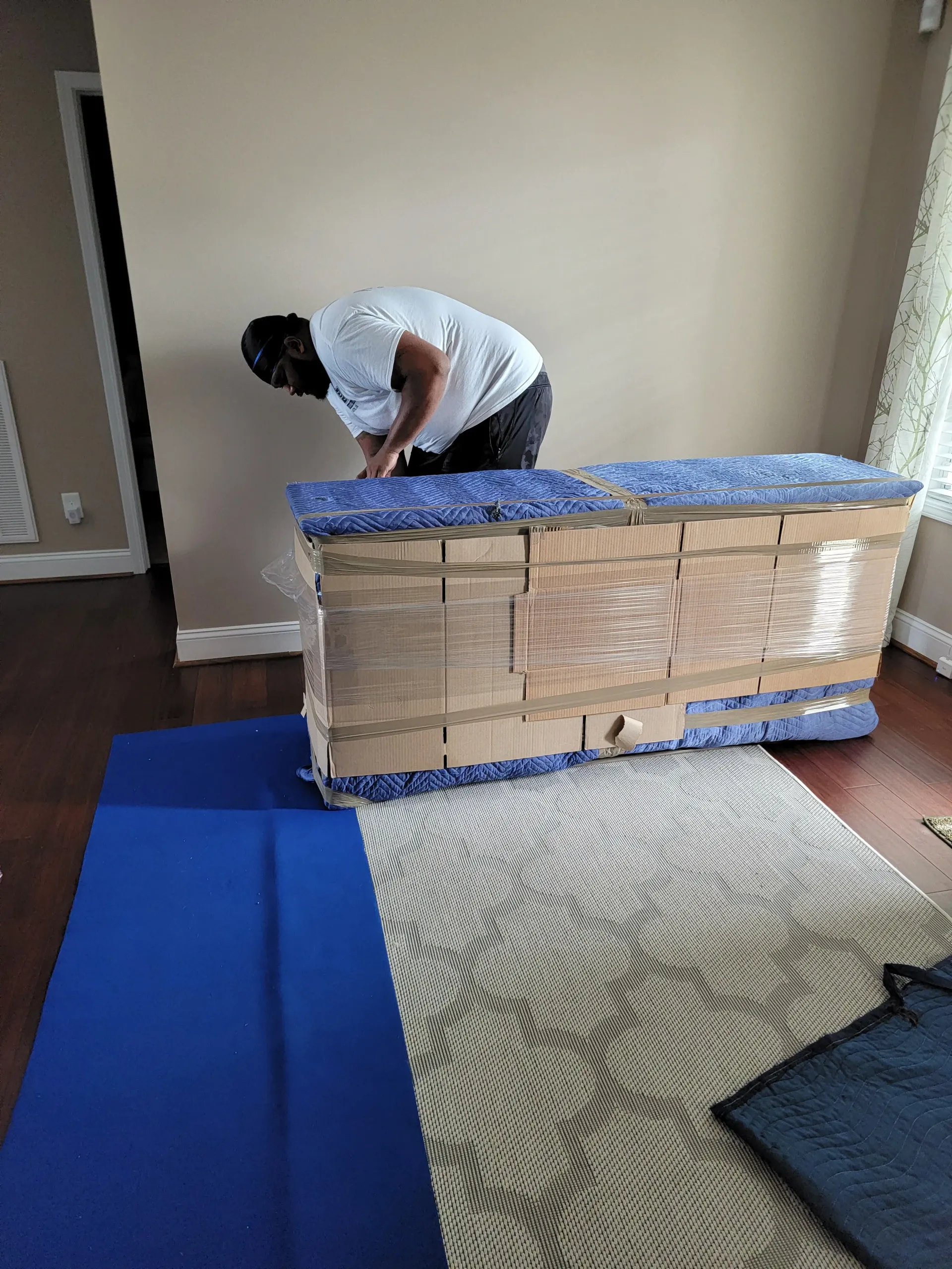 A person in a white shirt wrapping a large piece of furniture in protective blue padding and plastic film on a rug.