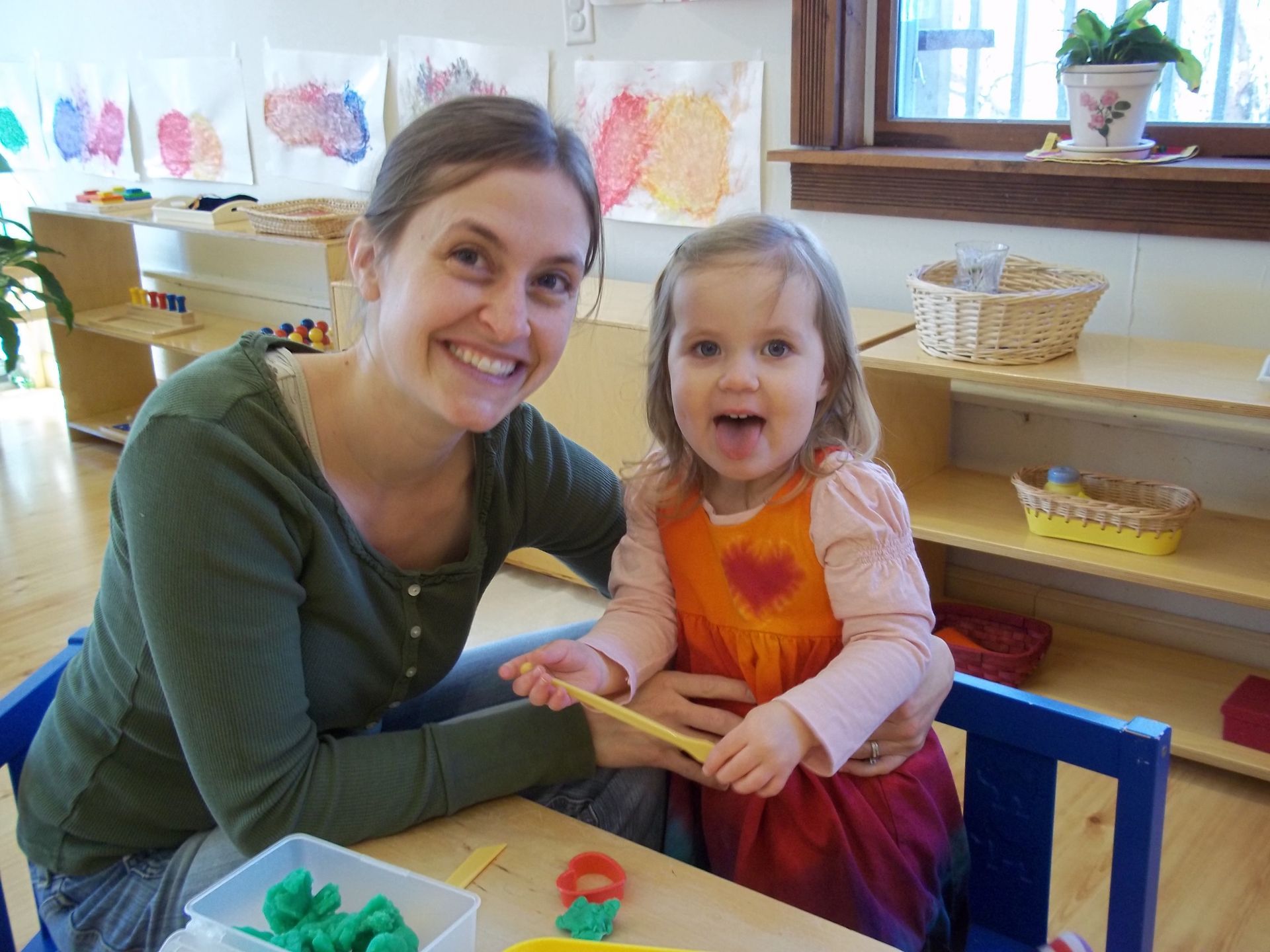 Montessori guide smiles with child. They're at a table with playdough, in a classroom with art on the wall.