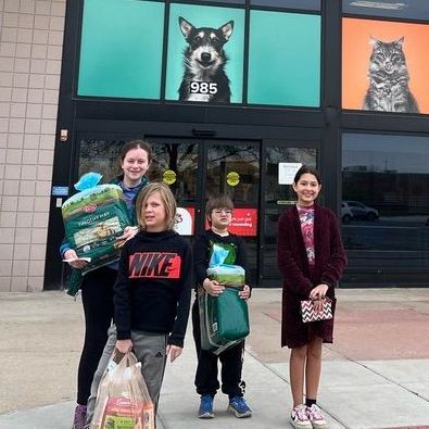 Four Montessori children stand outside a store, holding pet food. A pet shop is behind them with animal pictures.