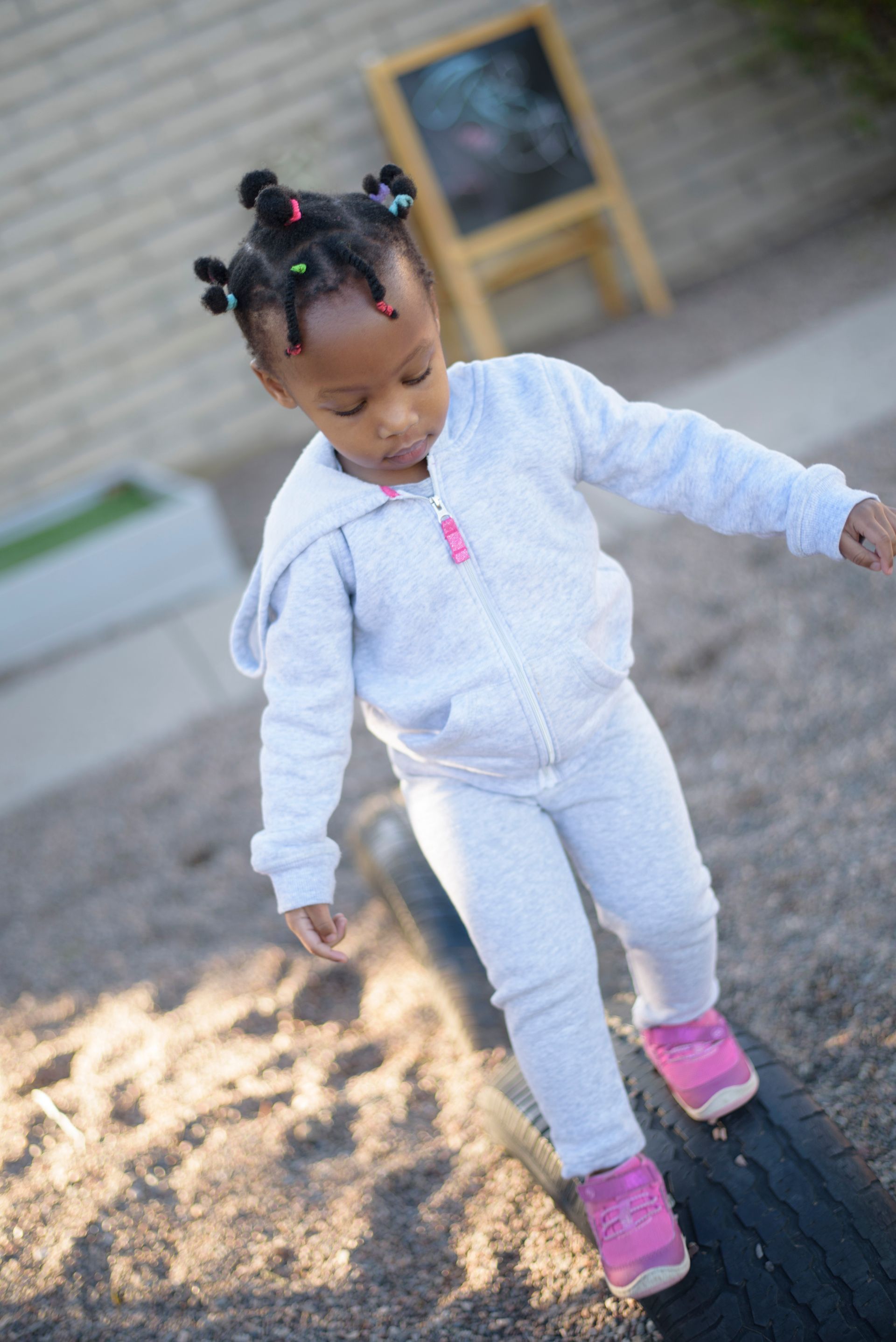 Child in a white tracksuit and pink shoes walking on a paved path outdoors.