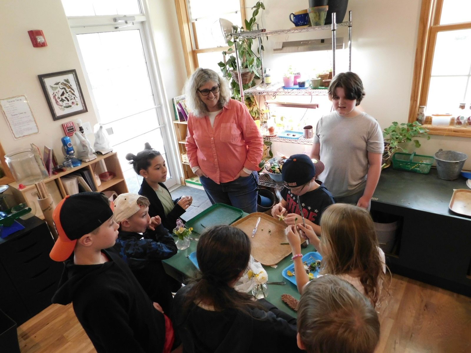 Montessori Children and a guide gather around a table, possibly examining items, inside a brightly lit room.