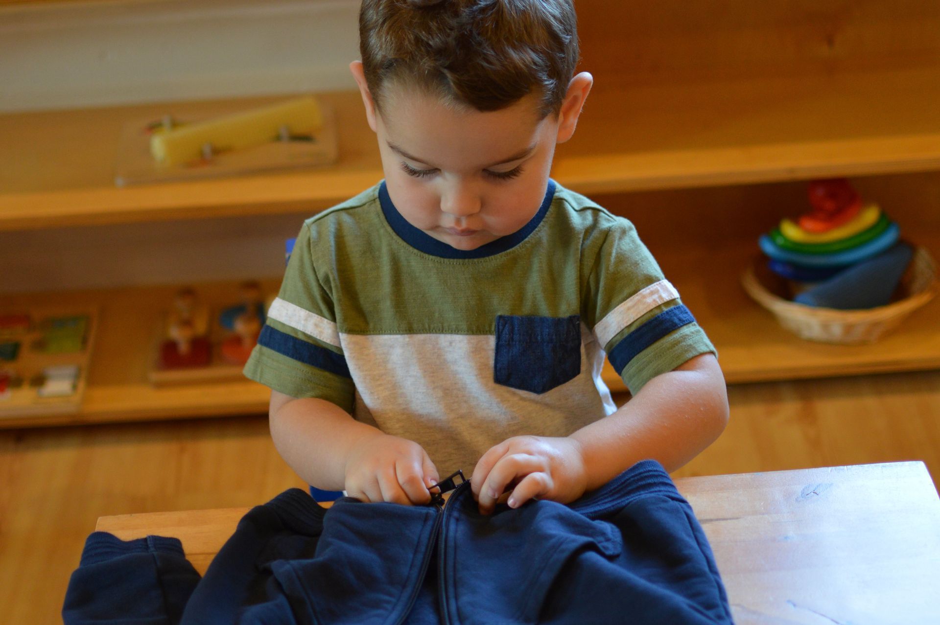 Montessori Child zipping a navy blue jacket at a wooden table in a room with shelves.