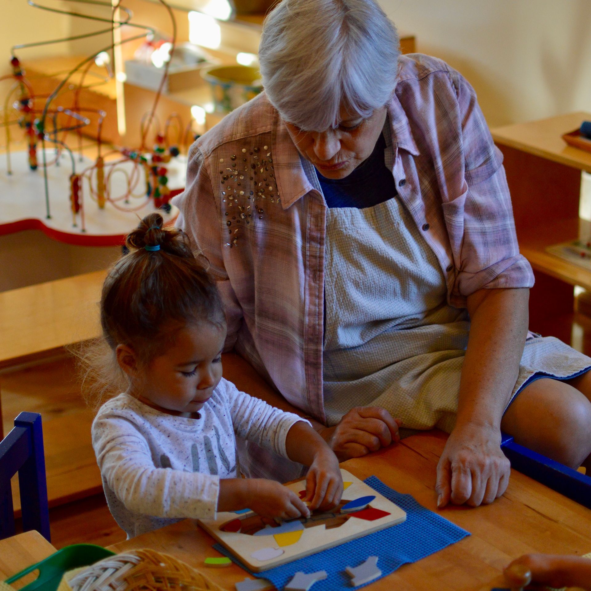 Montessori guide watches as a child works on a puzzle at a wooden table in a well-lit room.