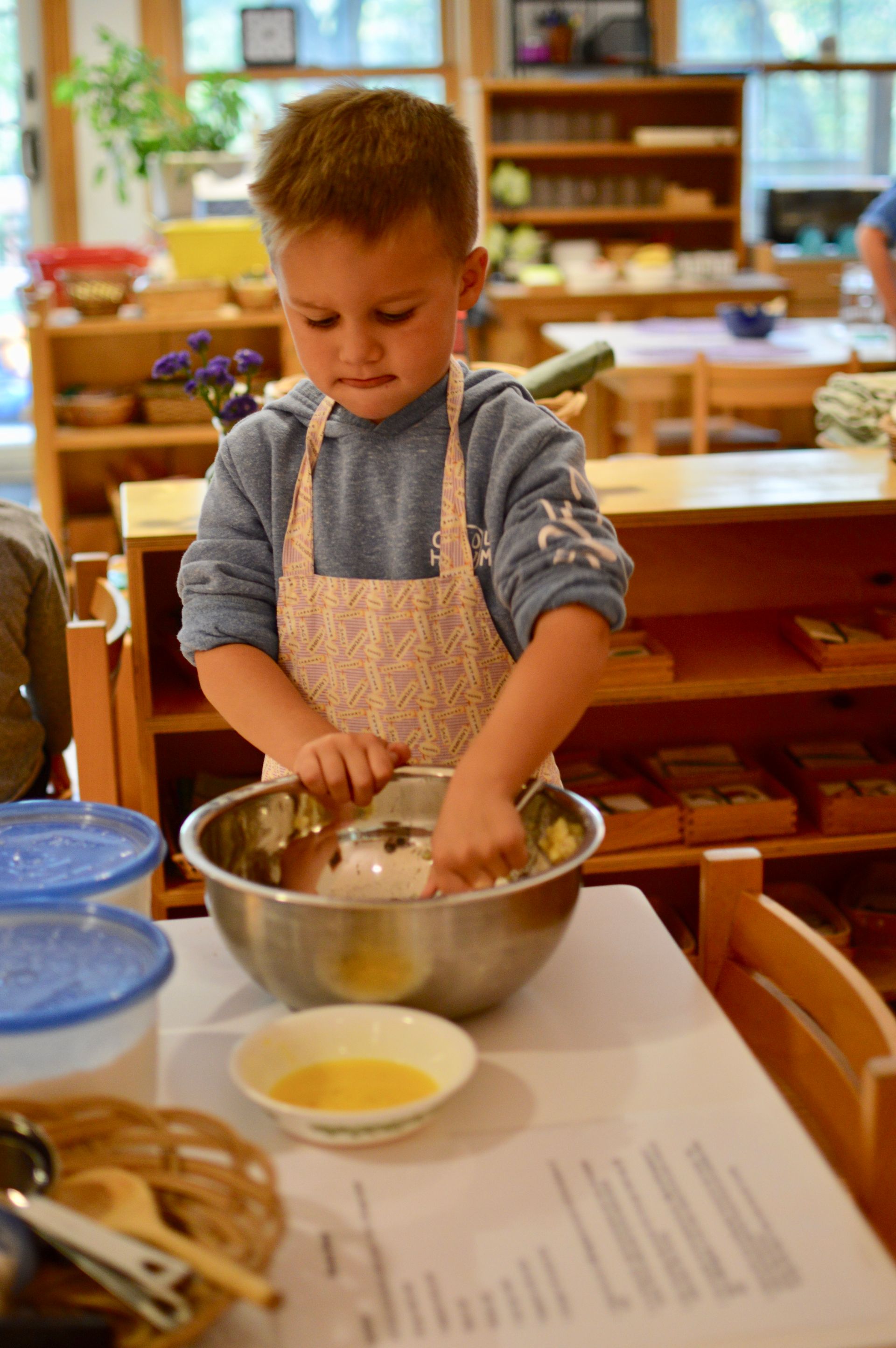 Montessori child in an apron mixing ingredients in a bowl on a table. He is focused, with a bright, sunny room in the background.