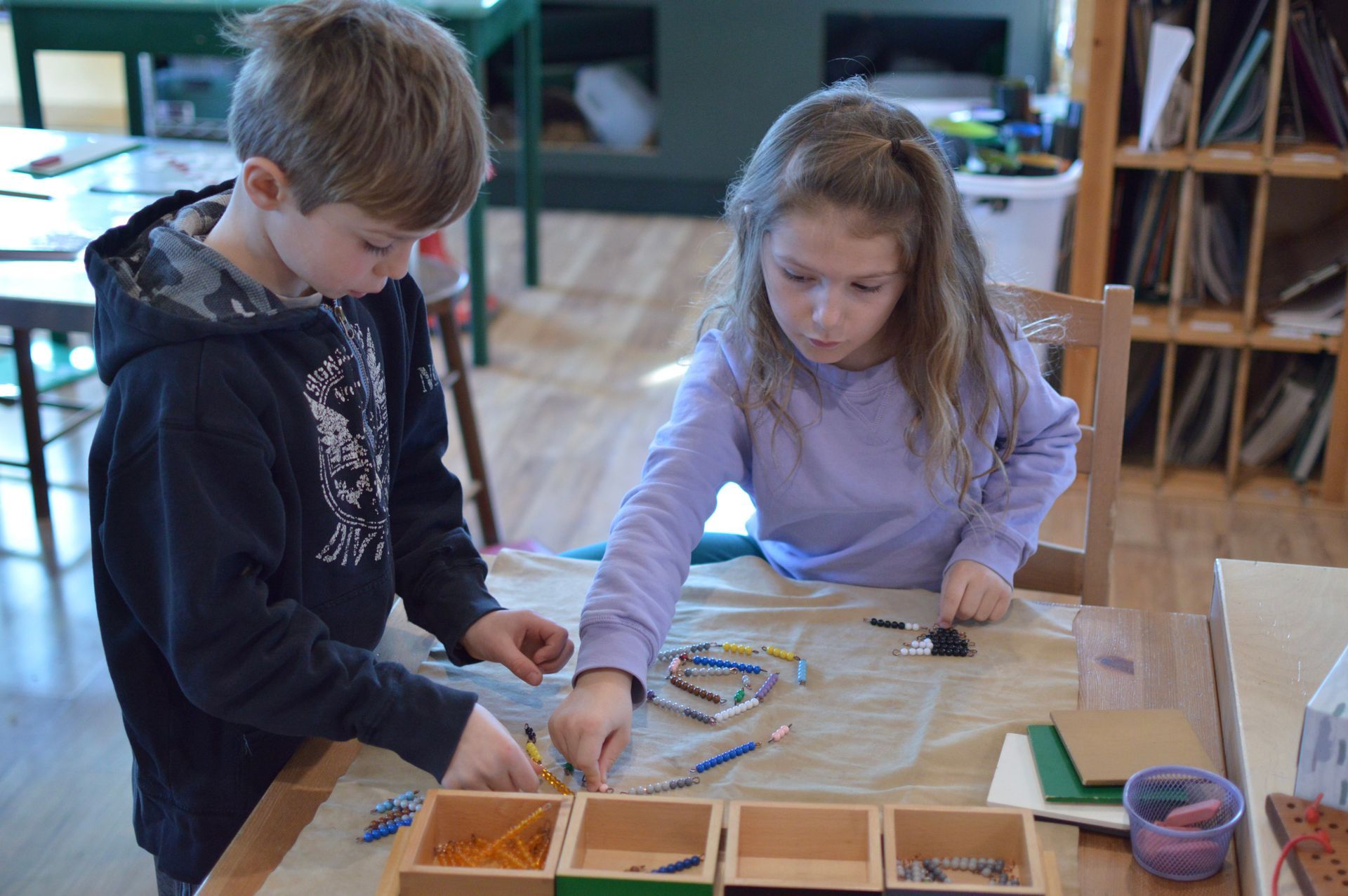 Two Montessori children working together at a table with small wooden boxes and objects, focused.