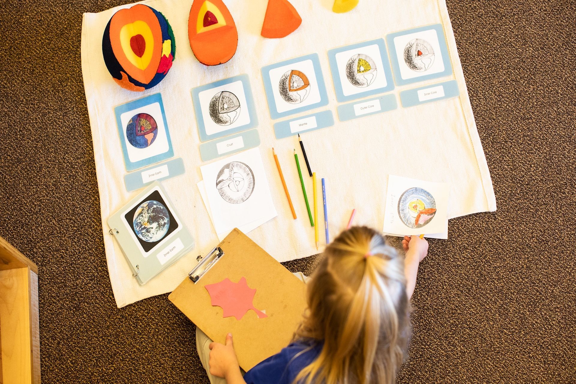 Child drawing on paper at a classroom table with art supplies and picture cards spread out.