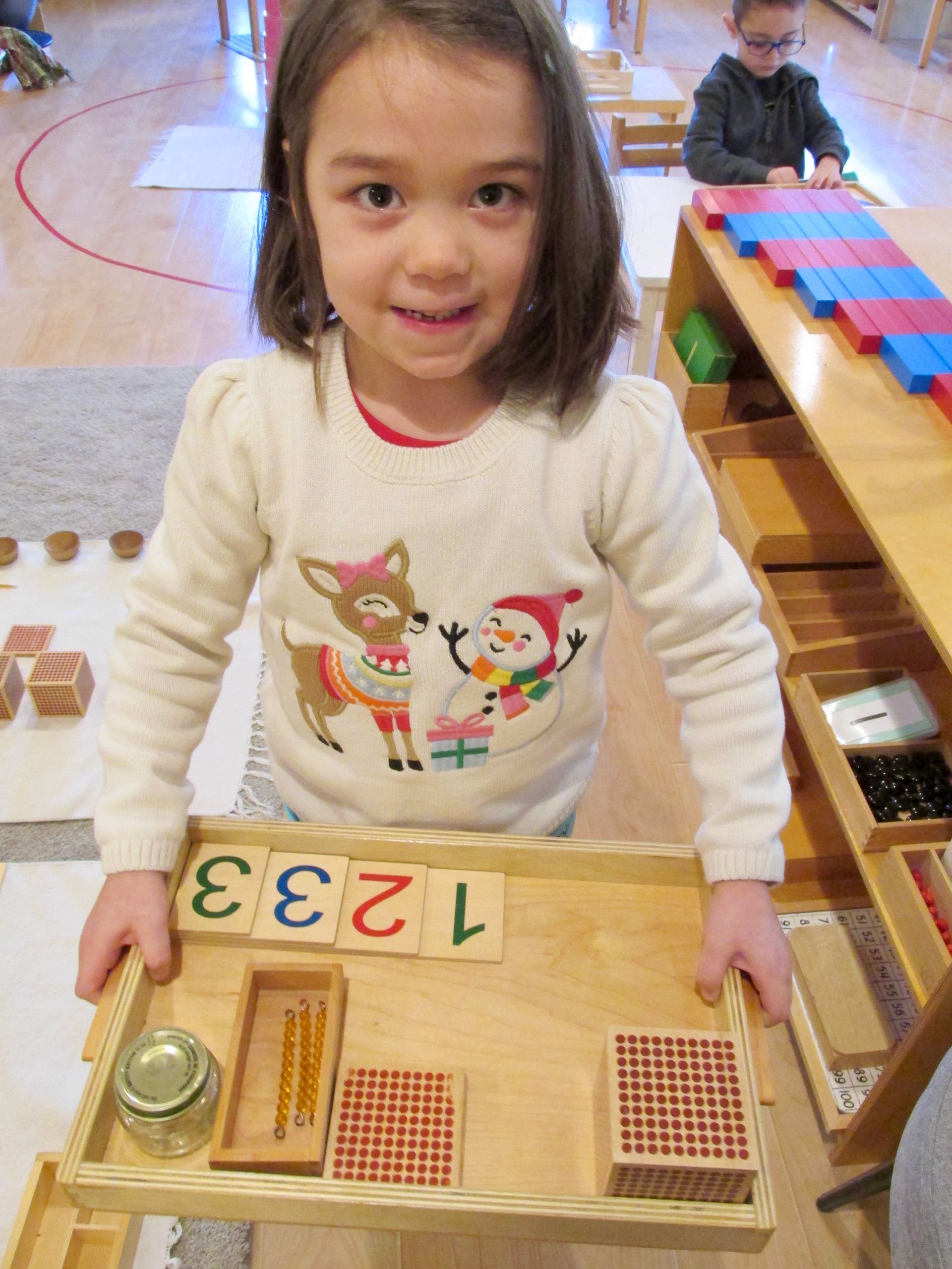 Montessori child holding tray with math materials, smiling in a Montessori classroom.