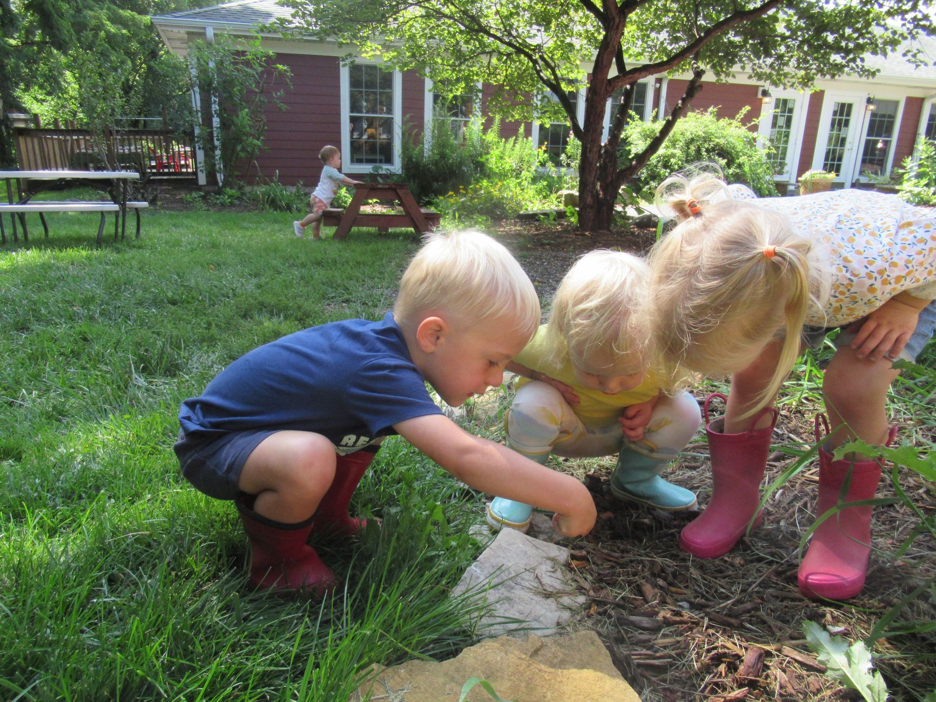 Three young Montessori children in rain boots crouch outdoors, examining soil.