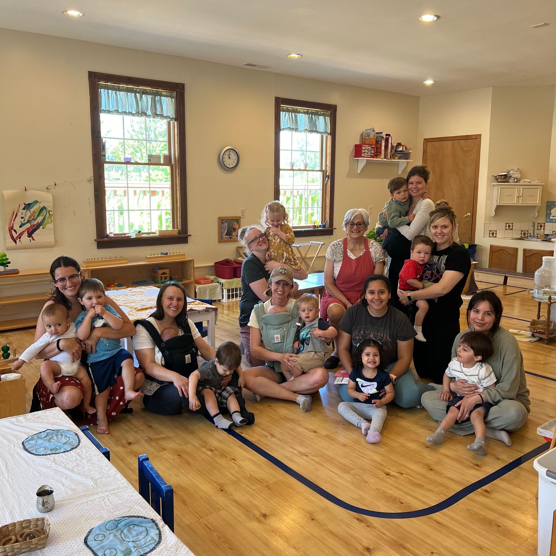 Group of adults and babies in a brightly lit room with wooden floors. Many are holding babies.