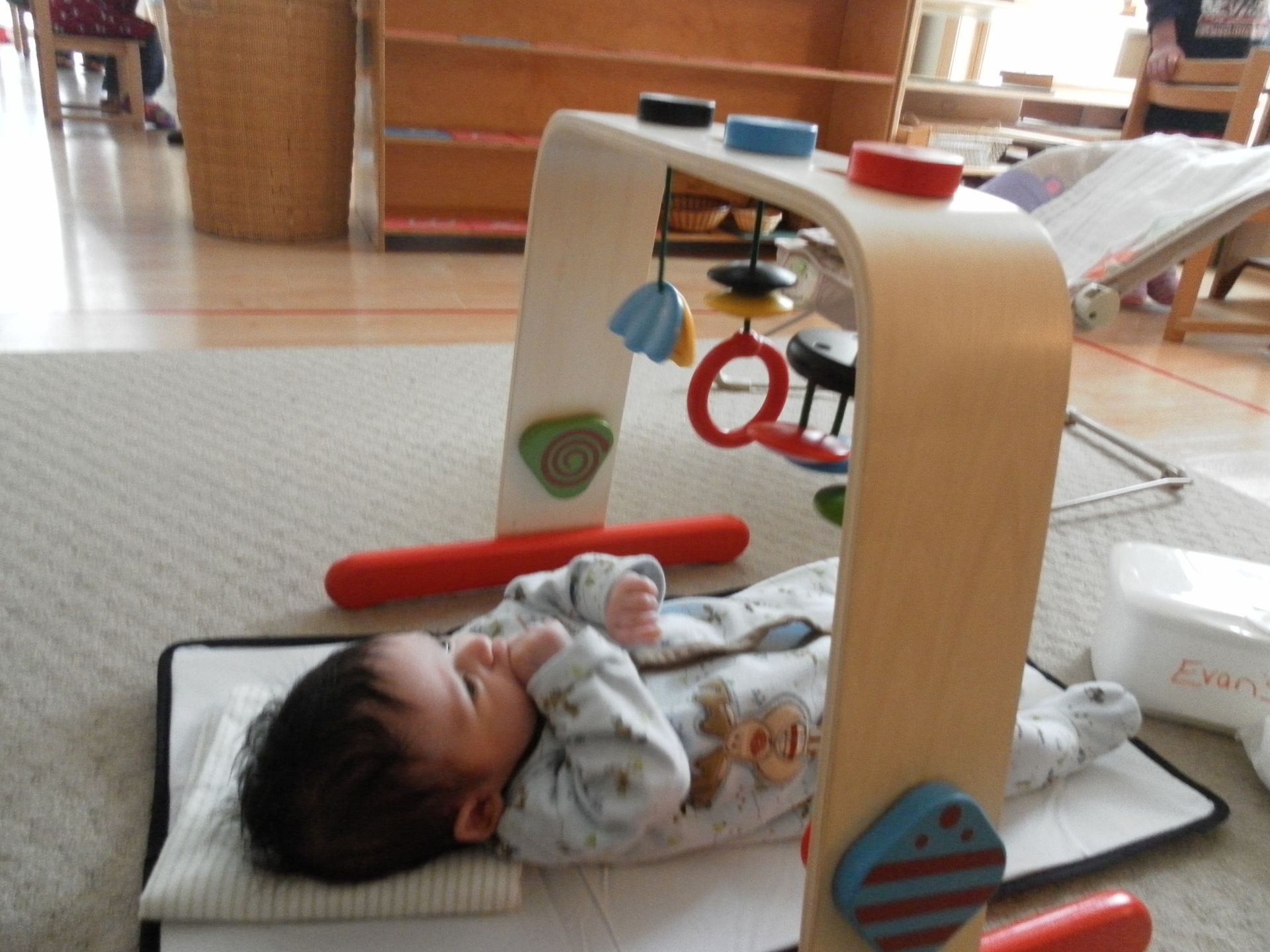 Montessori child lying on mat, looking at activity gym with hanging toys. Indoors on a carpeted floor.
