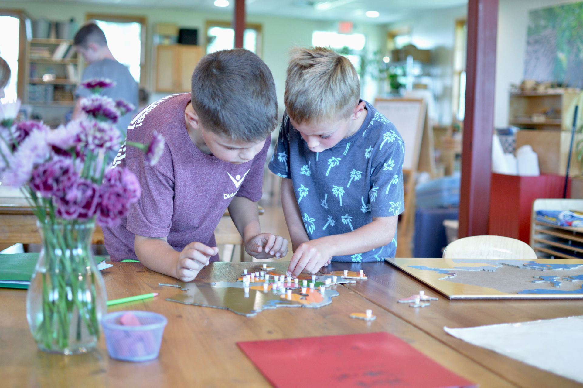 Two children lean over a table, carefully building with small pieces in a bright classroom.