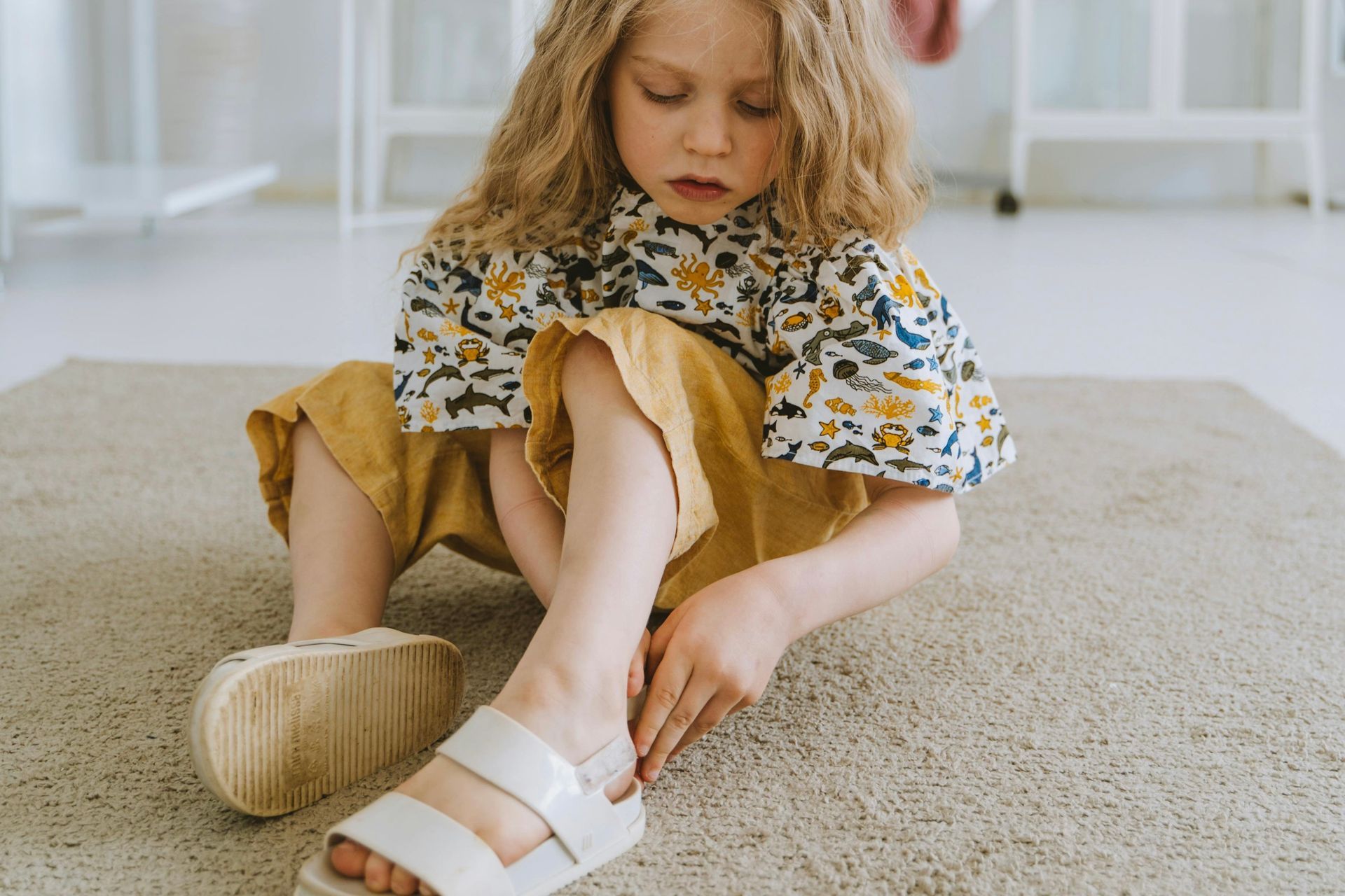 Child sitting on a carpet, fastening a white sandal while wearing a floral top and mustard pants