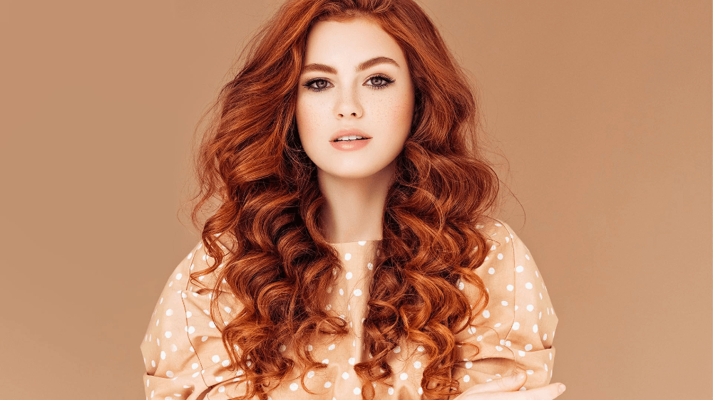 Vrouw met lang, krullend rood haar, gekleed in een beige blouse met stippen, tegen een lichtbruine achtergrond.