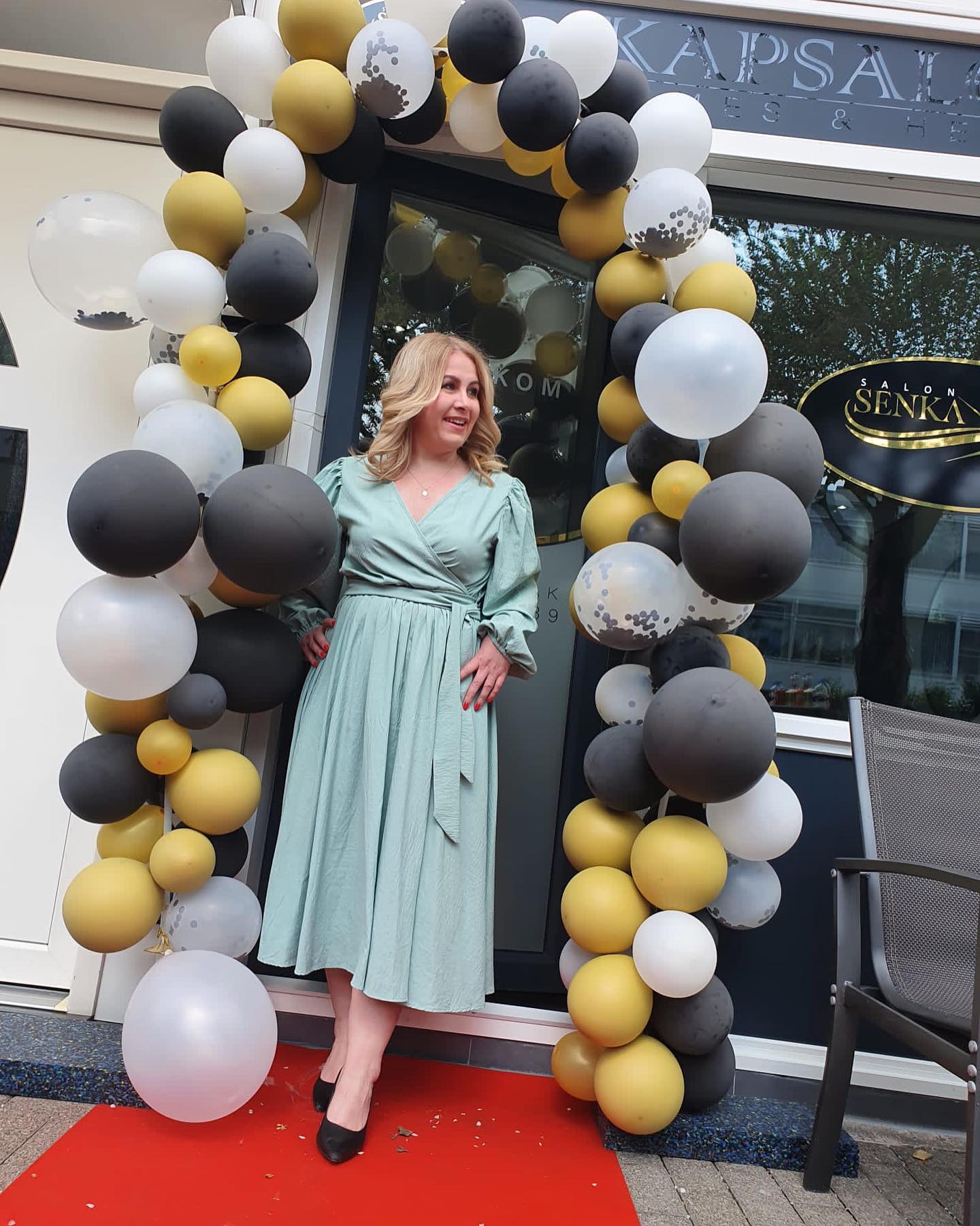Een vrouw in een turquoise jurk staat voor een winkelpui die versierd is met een ballonnenboog.
