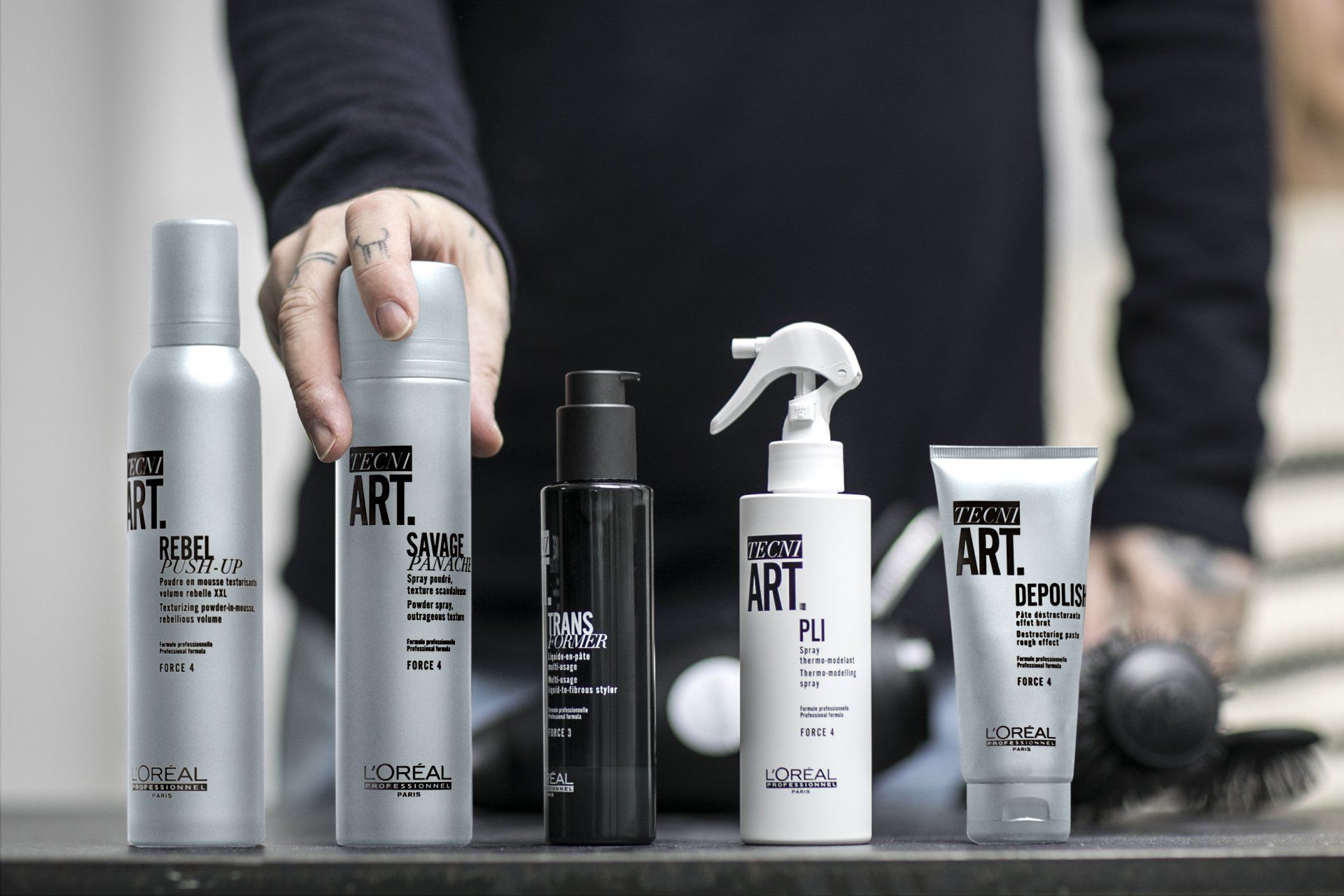 Een persoon houdt haarproducten vast: spray, schuim, gel en crème op een toonbank. De producten zijn zilverkleurig en zwart.