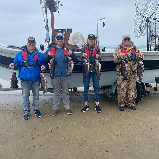 Four people holding up fish on a beach by a boat. Cloudy sky, wet sand.