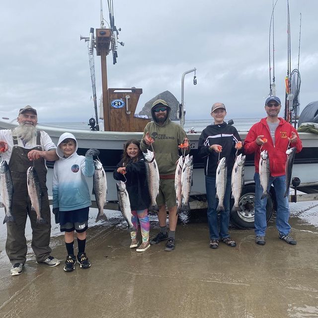 Group of people posing with caught fish on a dock near a fishing boat. Cloudy day.