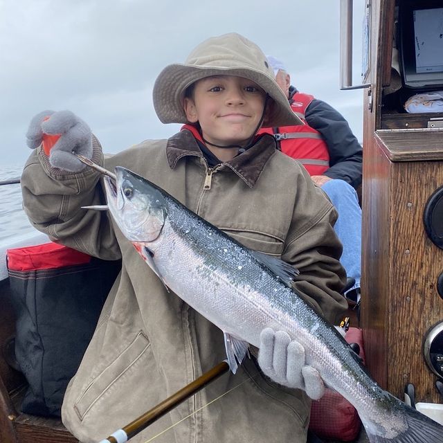 Boy in hat holds up a silver fish on a boat, smiling.