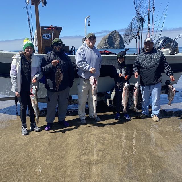 Group of people on a boat holding up fish they caught on a beach, Haystack Rock in the distance.