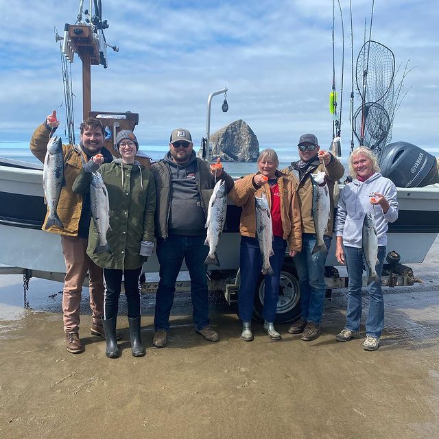 Group of people with fish next to a boat on a beach. People smiling and holding up their catch. Cloudy sky.
