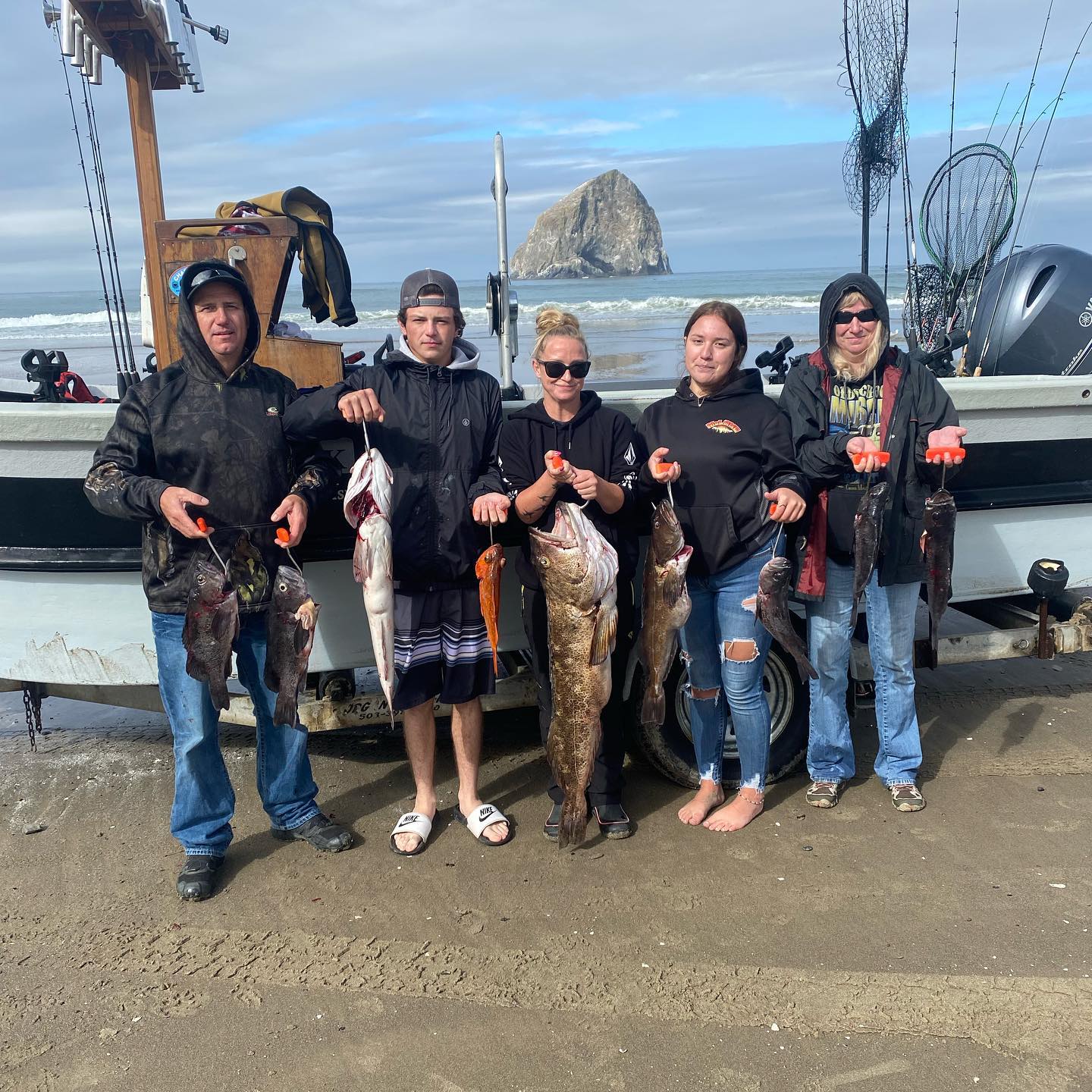 Five people holding fish they caught on a boat by the ocean; Haystack Rock in background.