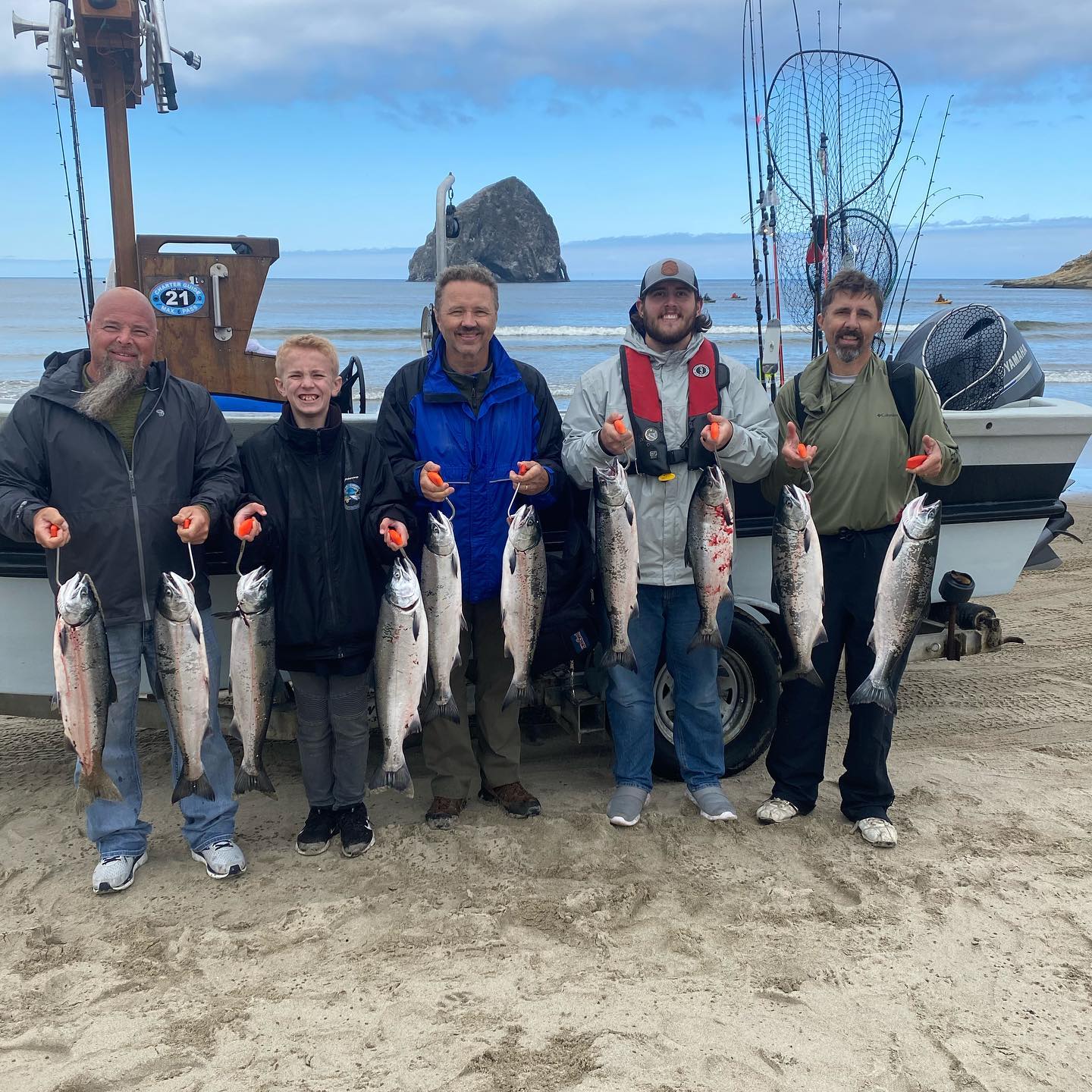 Five people on a beach hold up caught fish; a boat and ocean in the background.