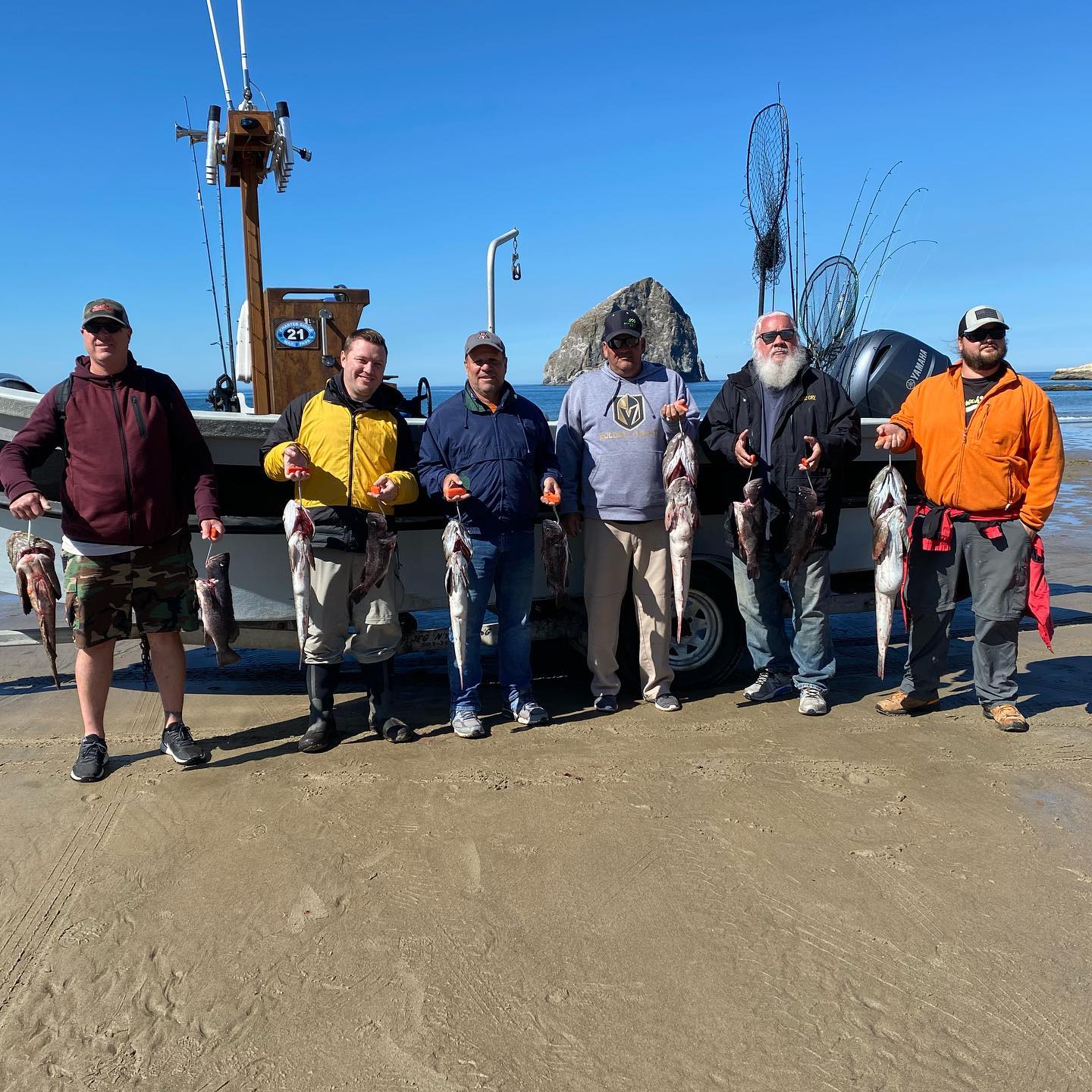 Group of people holding up fish they caught by the shore on a sunny day. Boat in background.