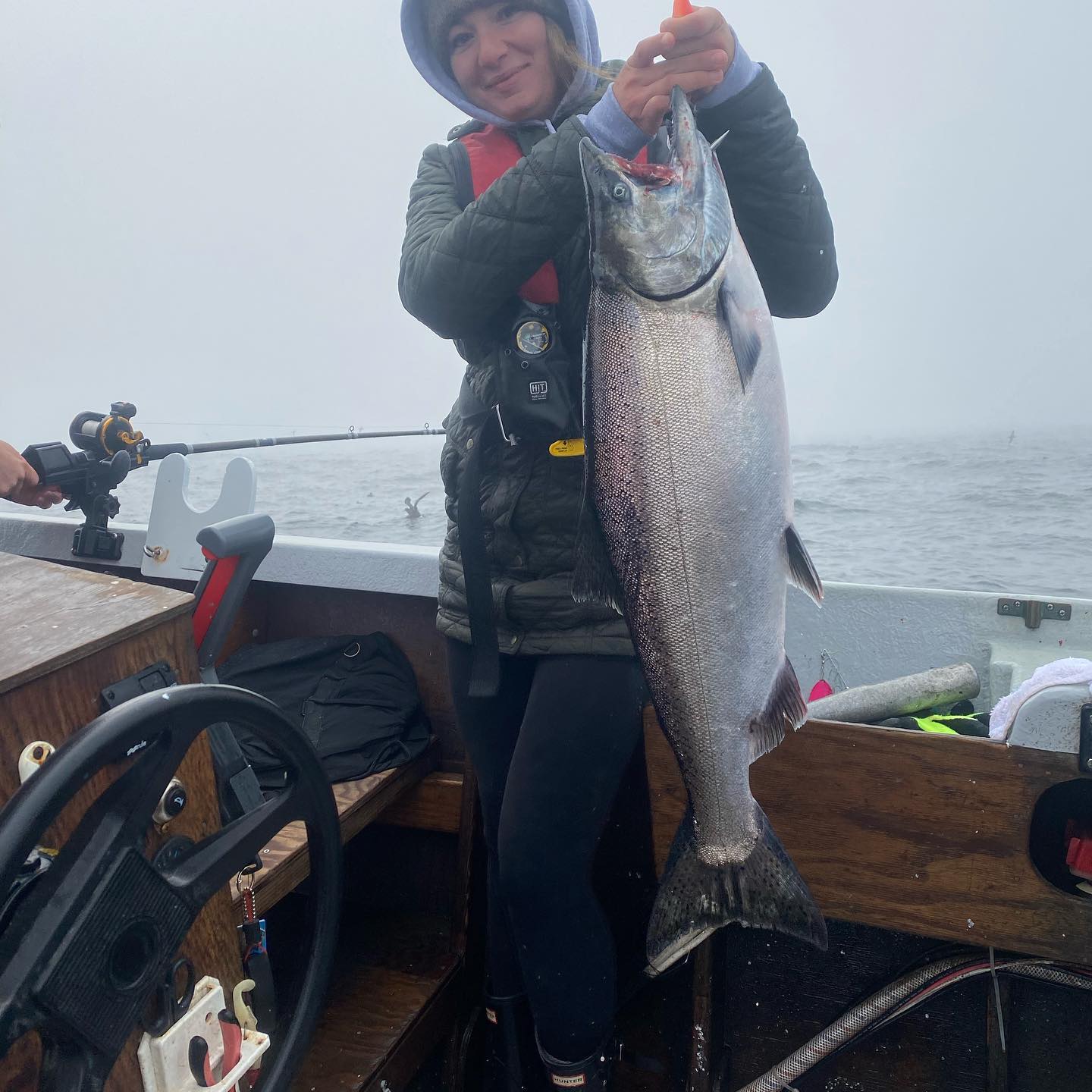 Woman on a boat holds up a large silver fish, smiling. Overcast day, fishing rod visible.