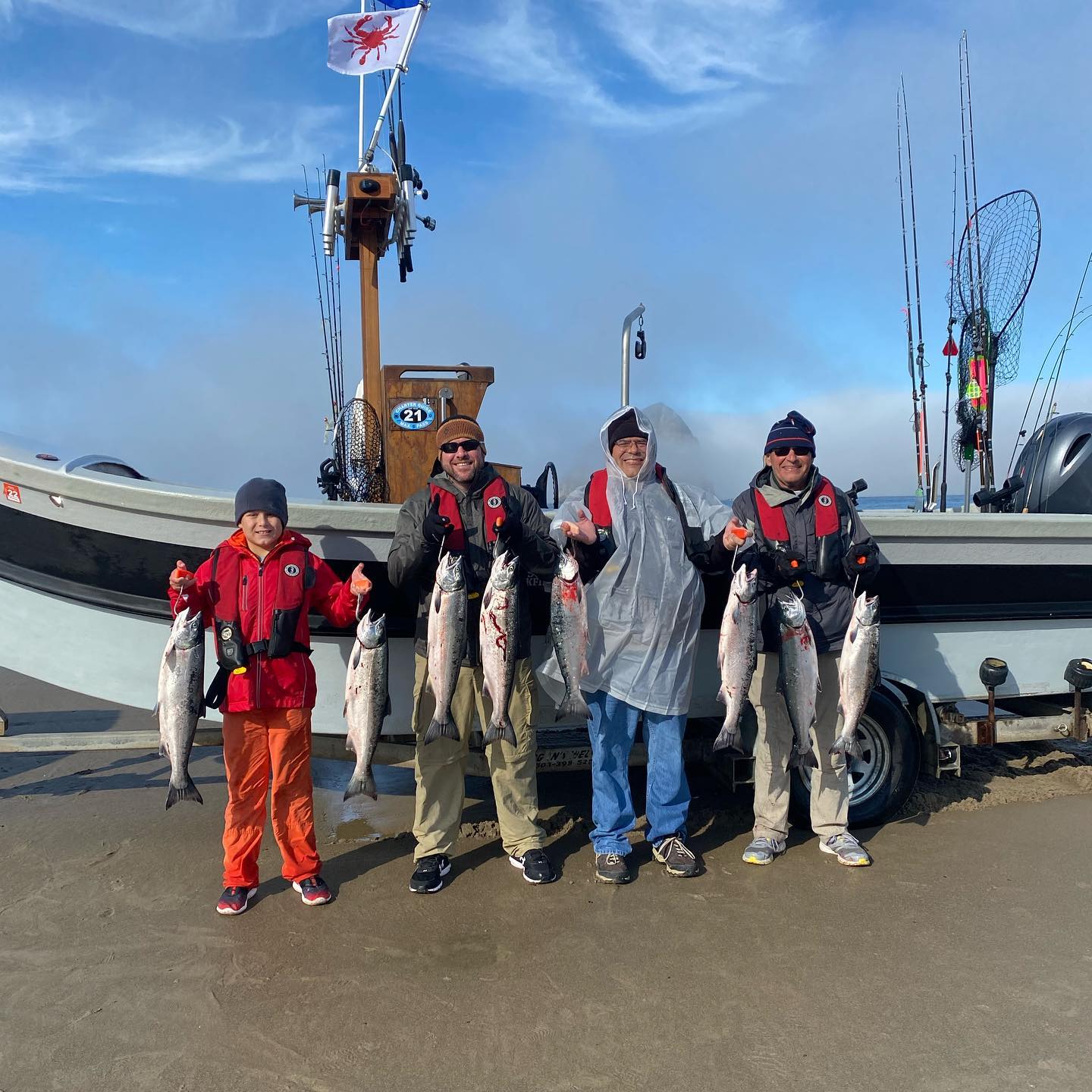 Four people hold fish, smiling in front of a boat with a crab flag, sunny outdoor setting.