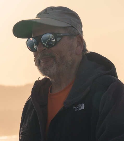 Man in sunglasses and cap, wearing a dark jacket, looking towards the sun.
