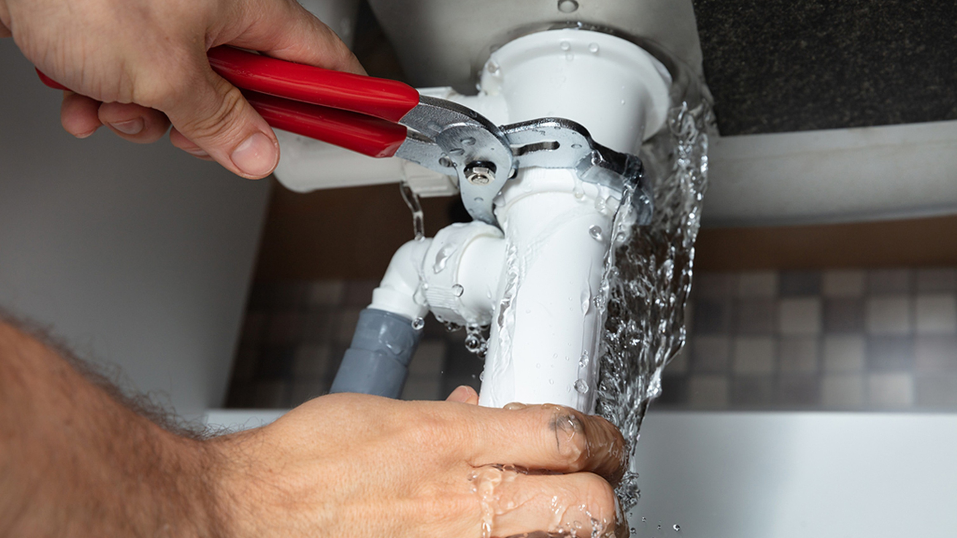 Person using pliers to fix a leaking white sink drain pipe, water spraying.