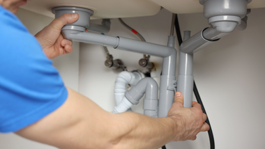 Hands assembling gray plumbing pipes under a white sink.