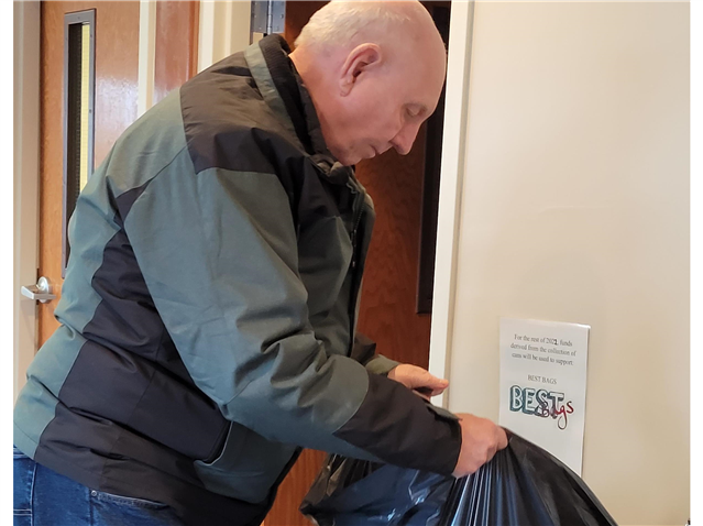 Mike Deshon is putting pop cans in a trash bag next to a sign that says best bags.
