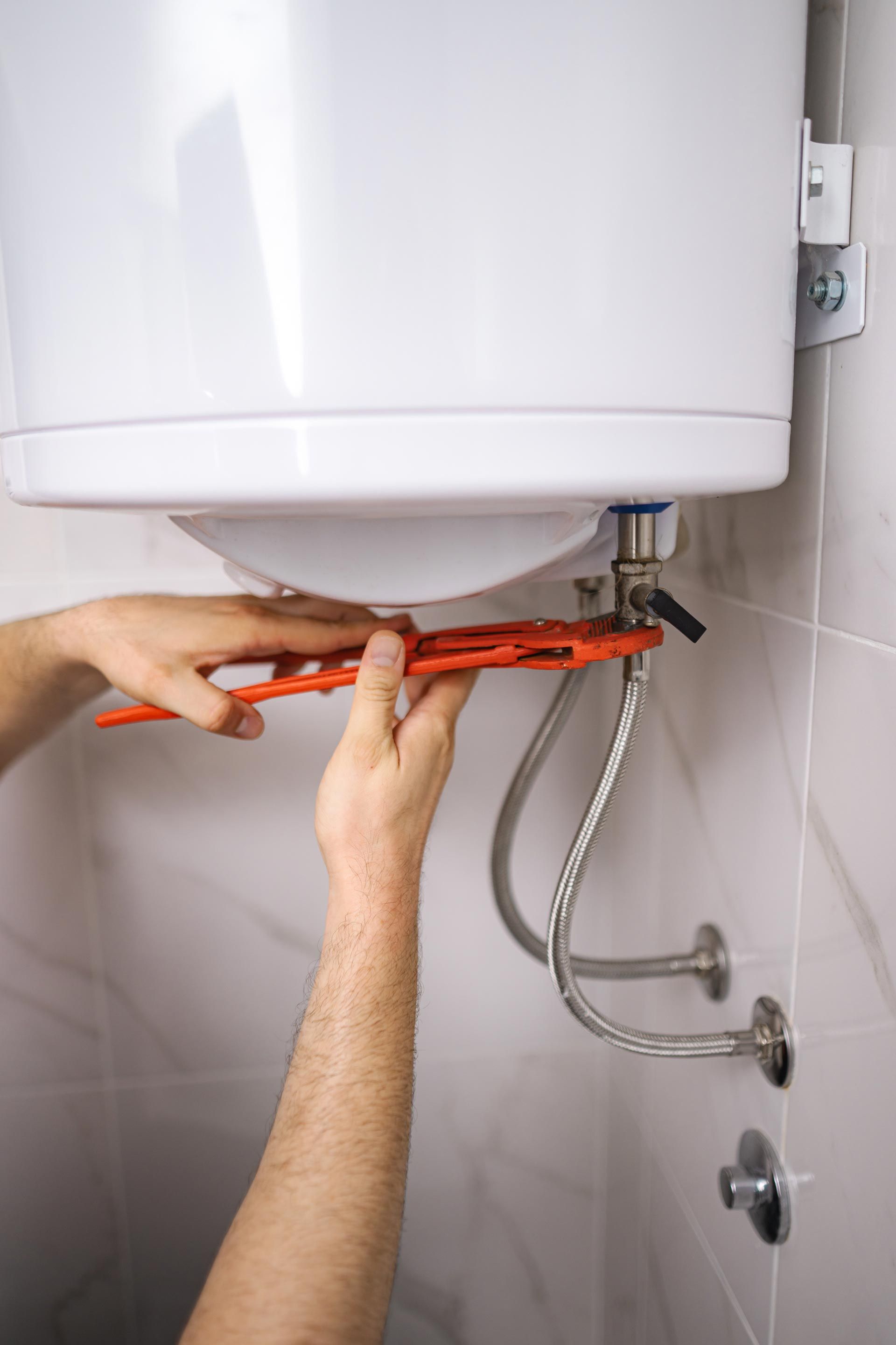 Technician tightening plumbing under a white water heater with an orange wrench in a tiled utility room
