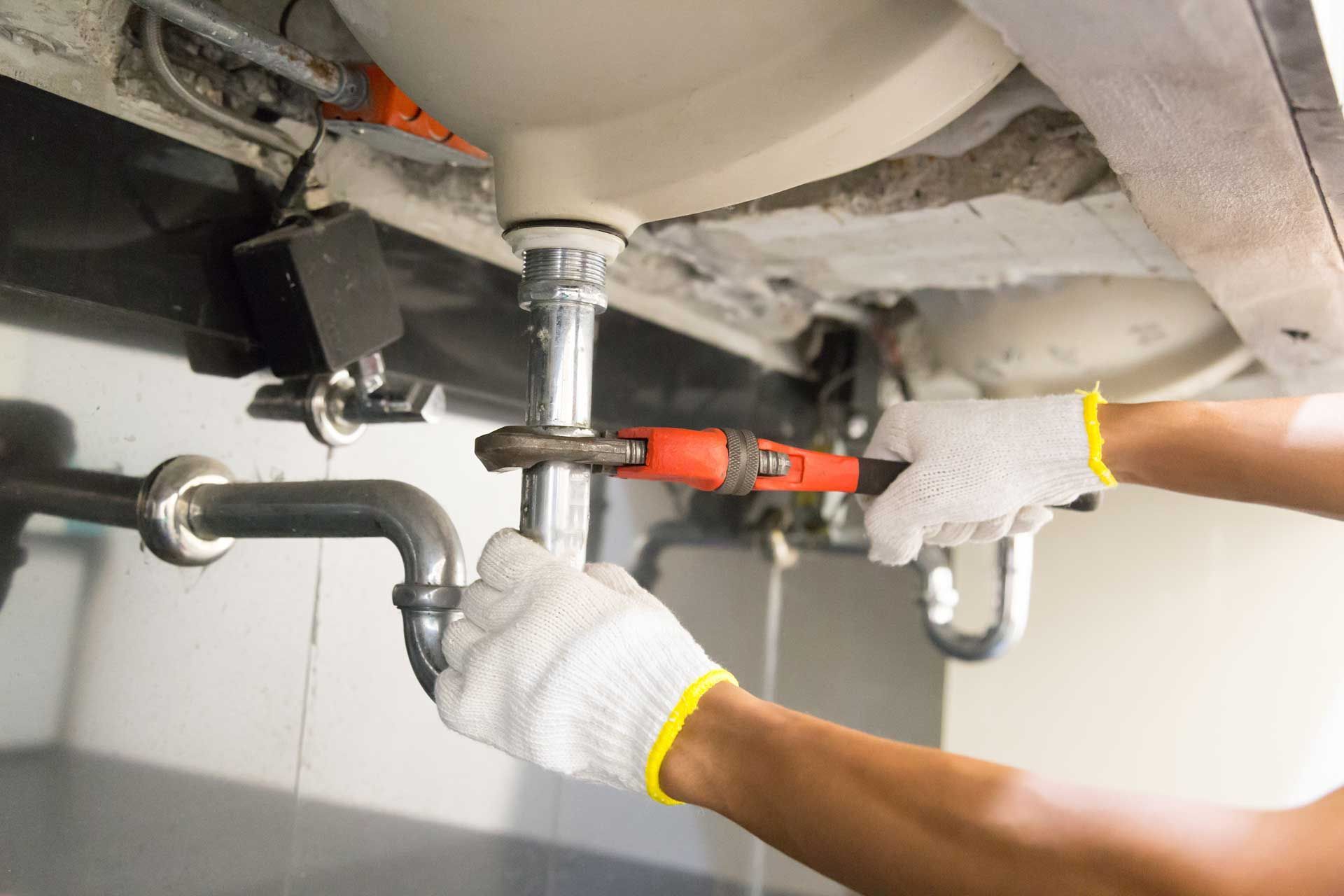 Technician using a wrench to fix plumbing under a sink, with exposed pipes and a red tool