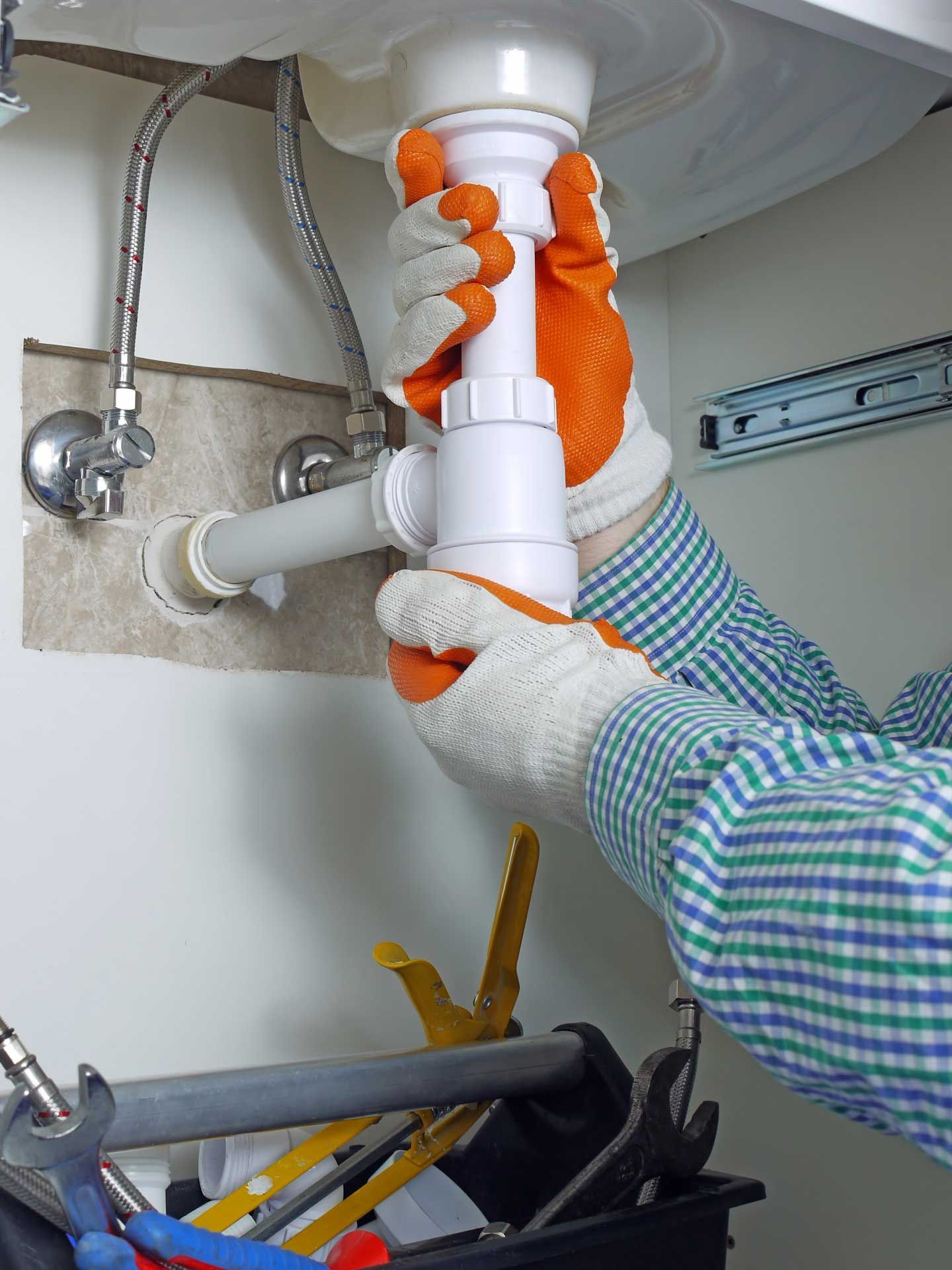 Hands wearing orange gloves assembling white PVC pipes under a kitchen sink.