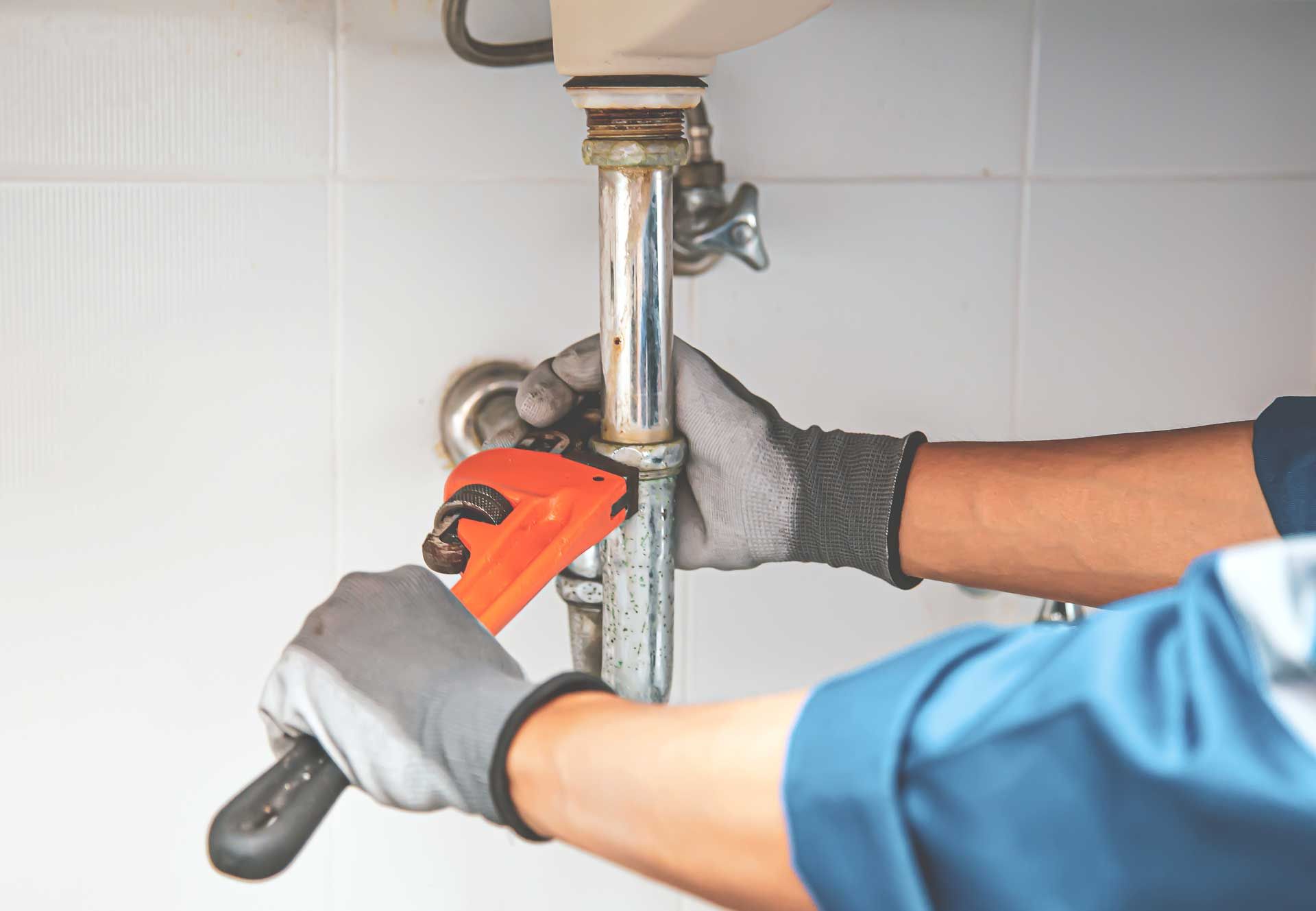 Plumber using an orange pipe wrench to loosen a chrome drain pipe under a sink