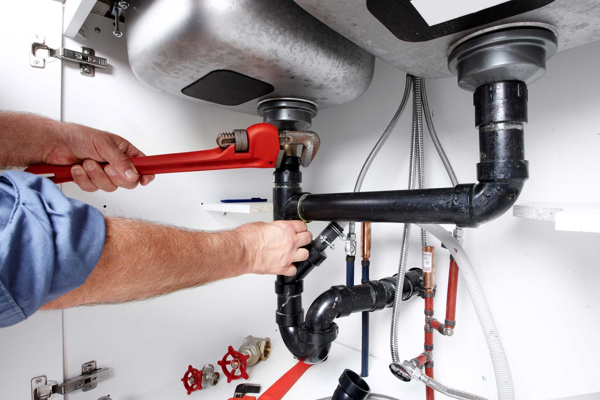 Plumber tightening black under-sink pipes with a red wrench beneath a kitchen sink