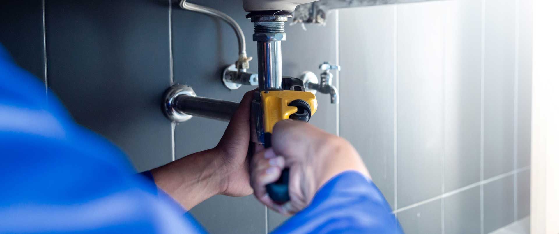 Person tightening a pipe valve with a wrench under a sink, wearing a blue jacket and gloves.